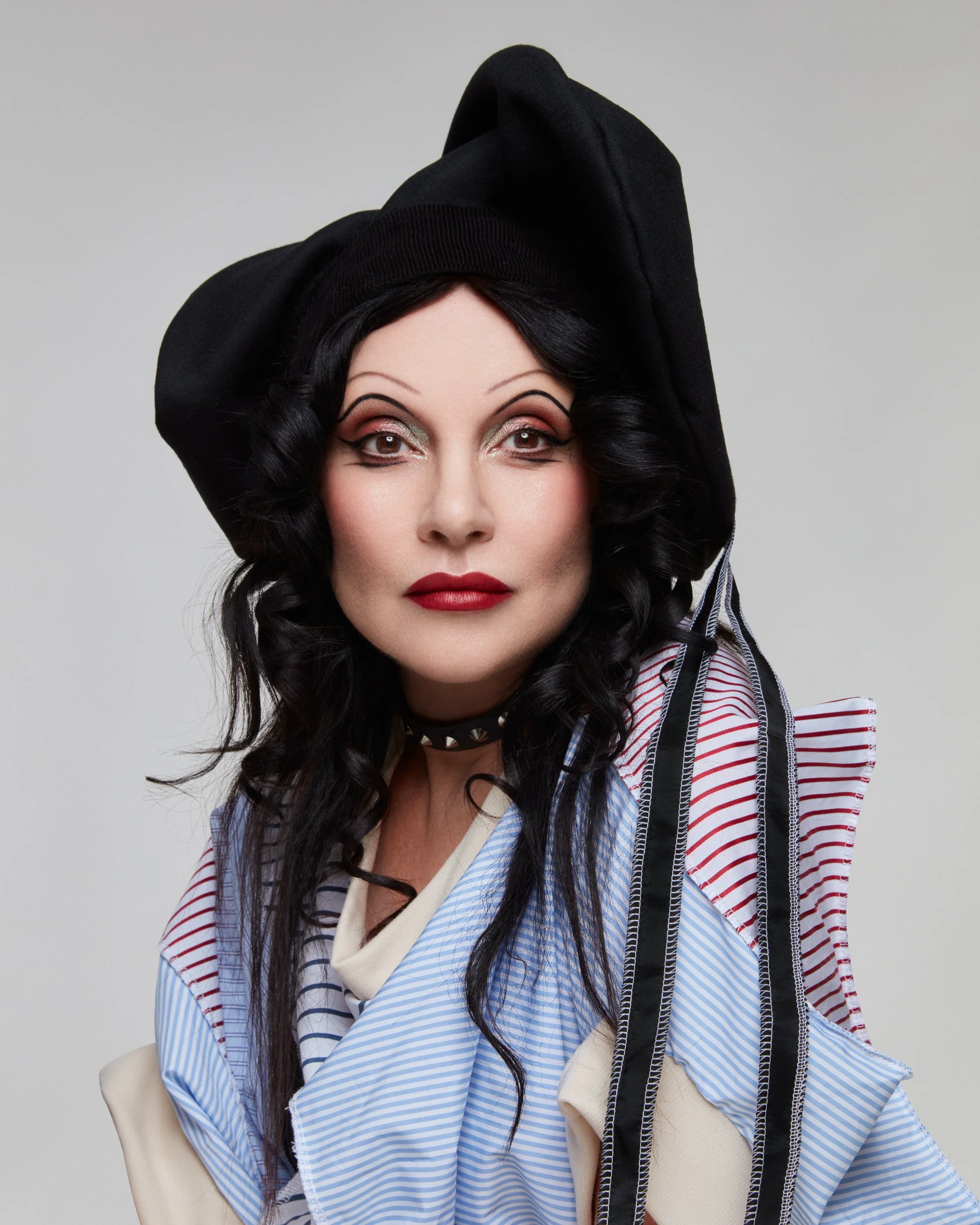 A woman with black curly hair and makeup, wearing a black headscarf, a striped shirt with ruffled shoulders, and a black choker.