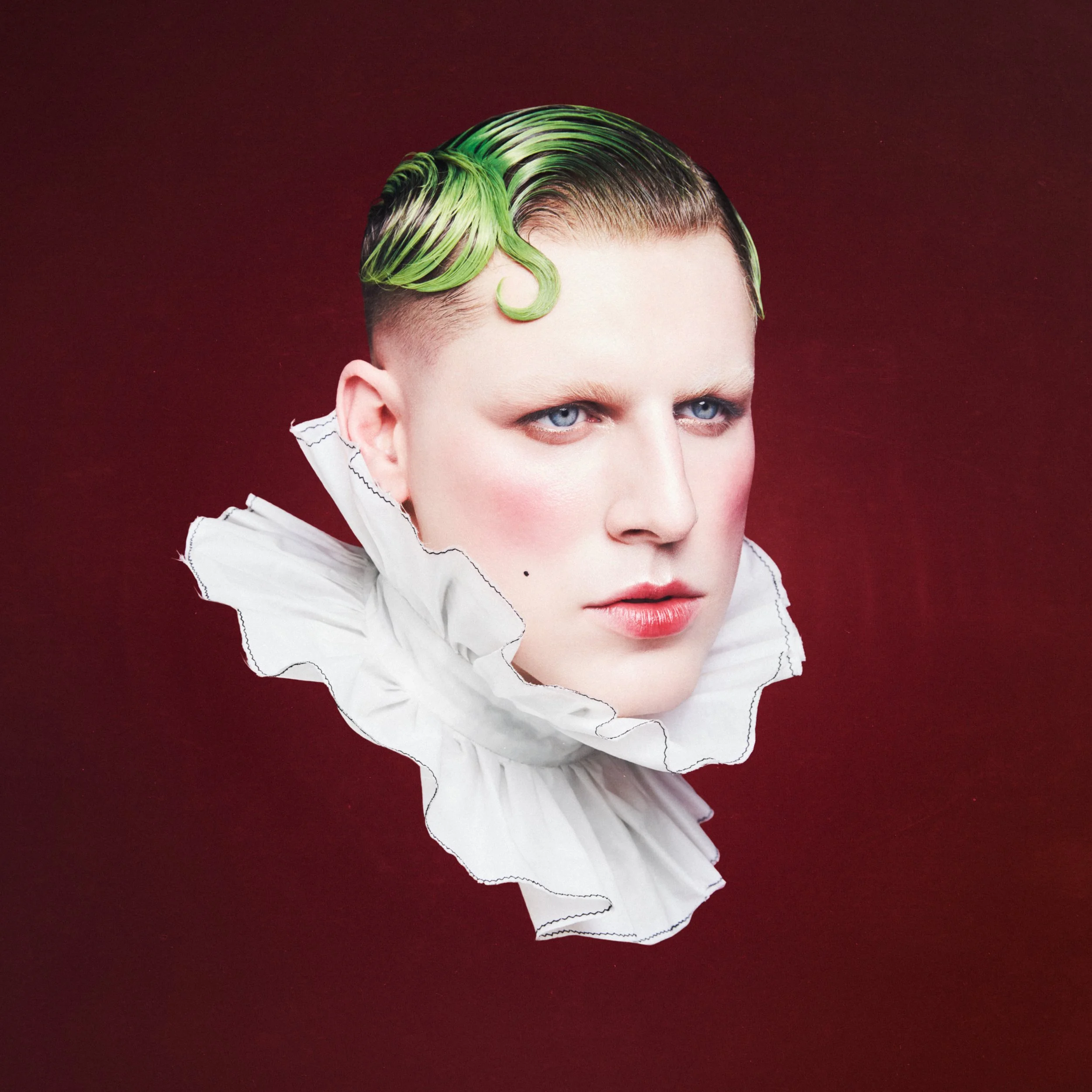 A person with short hair dyed green on top, gazing to the right, wearing a ruffled white collar against a dark red background.