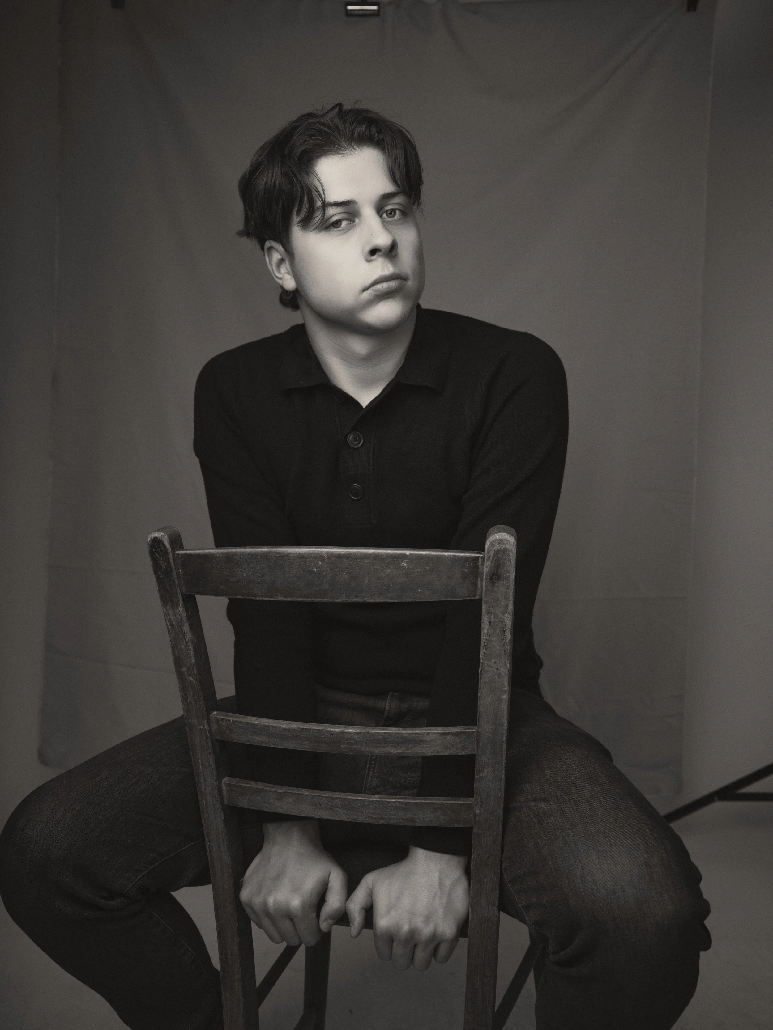 A young person with dark wavy hair, wearing a black polo shirt, sitting behind an old wooden chair, with a serious expression, in a plain gray room.