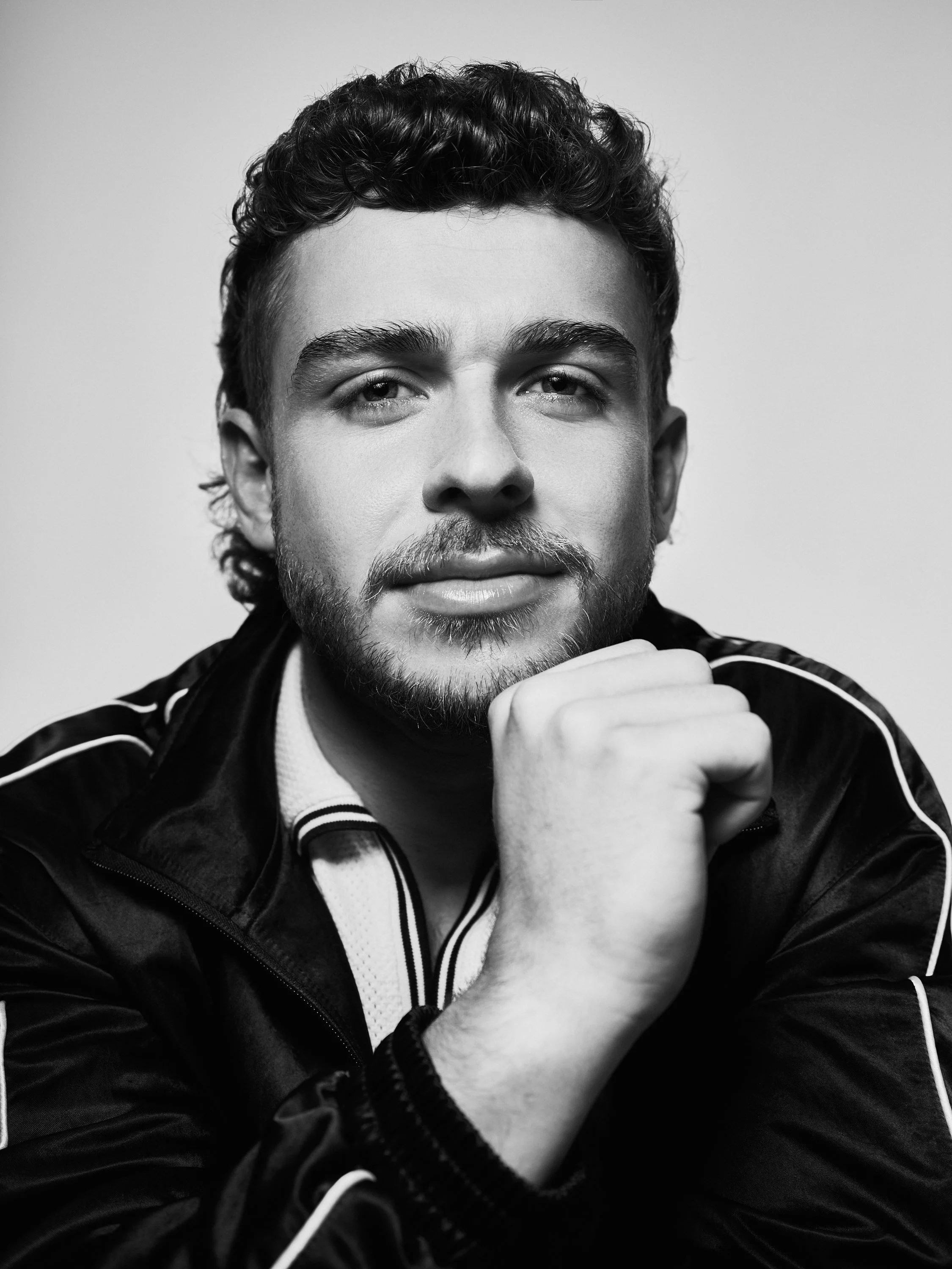 Black and white portrait of a young man with curly hair and a beard, resting his chin on his hand, wearing a jacket with white stripes.