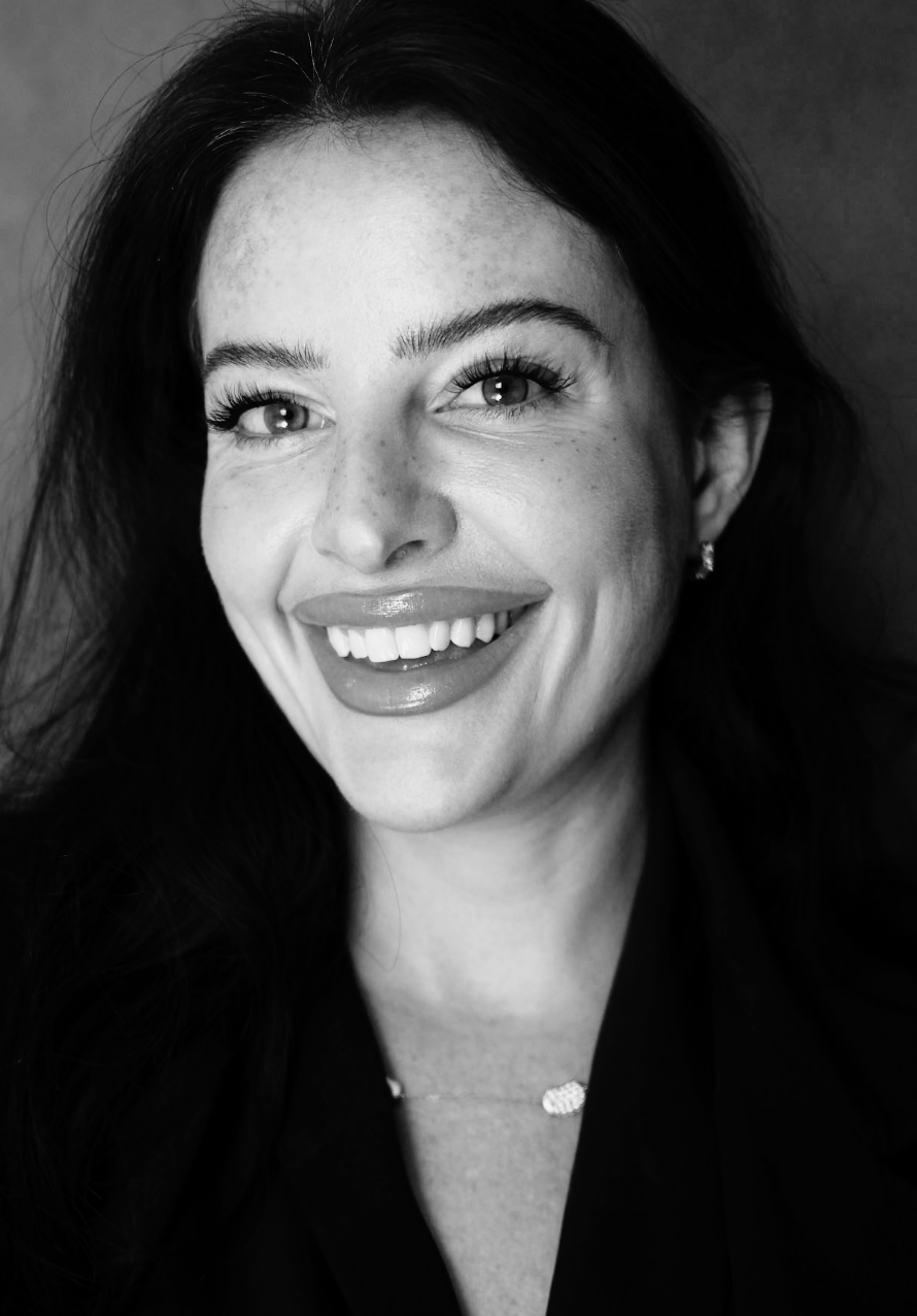 Black and white portrait of a smiling woman with long dark hair, bright eyes, and makeup, wearing a necklace and earrings.
