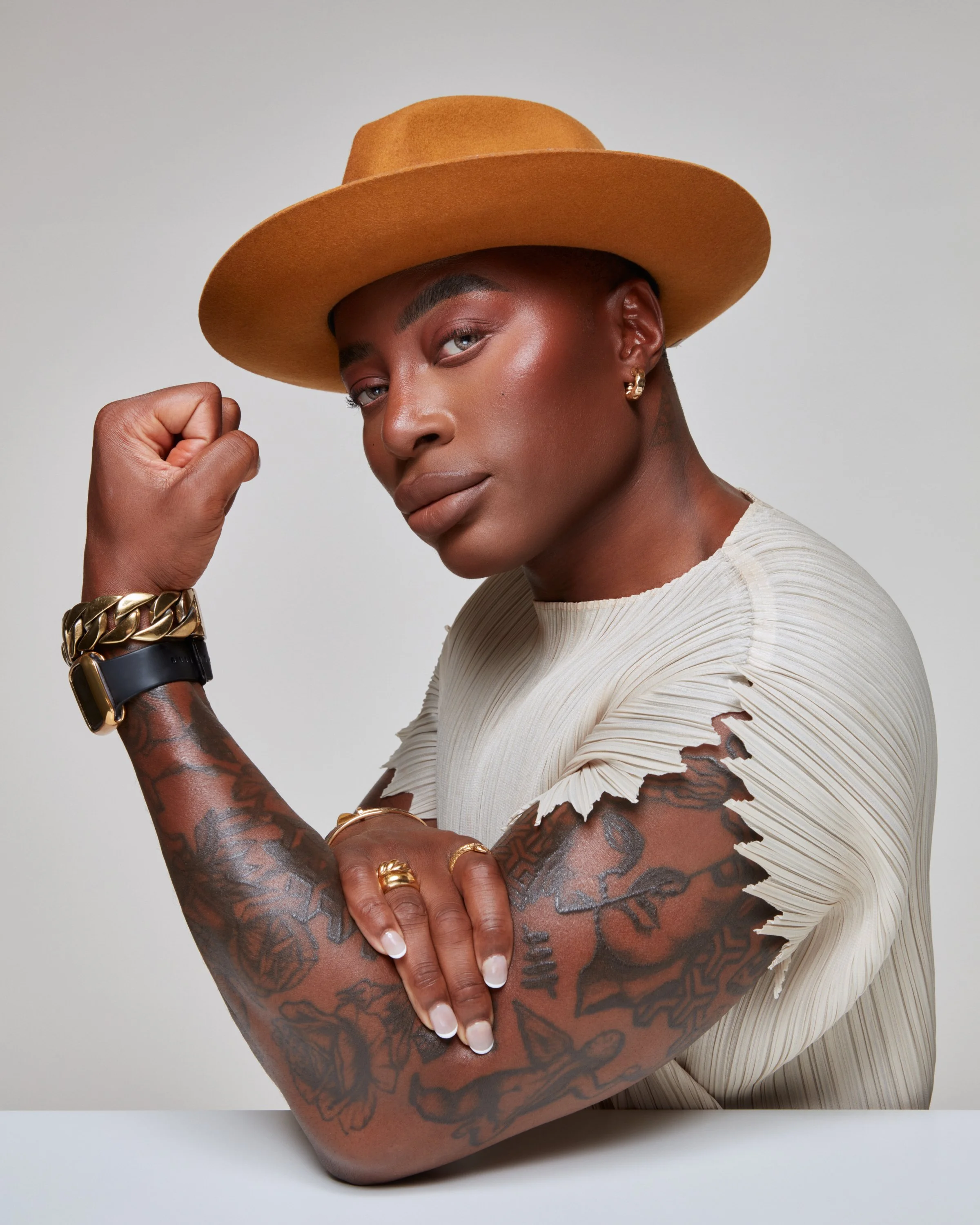 A woman with tattoos on her arms, wearing a tan hat and a white shirt with ripped sleeves. She is flexing her right arm, showing her tattoos, and has jewelry including rings, bracelets, and earrings.