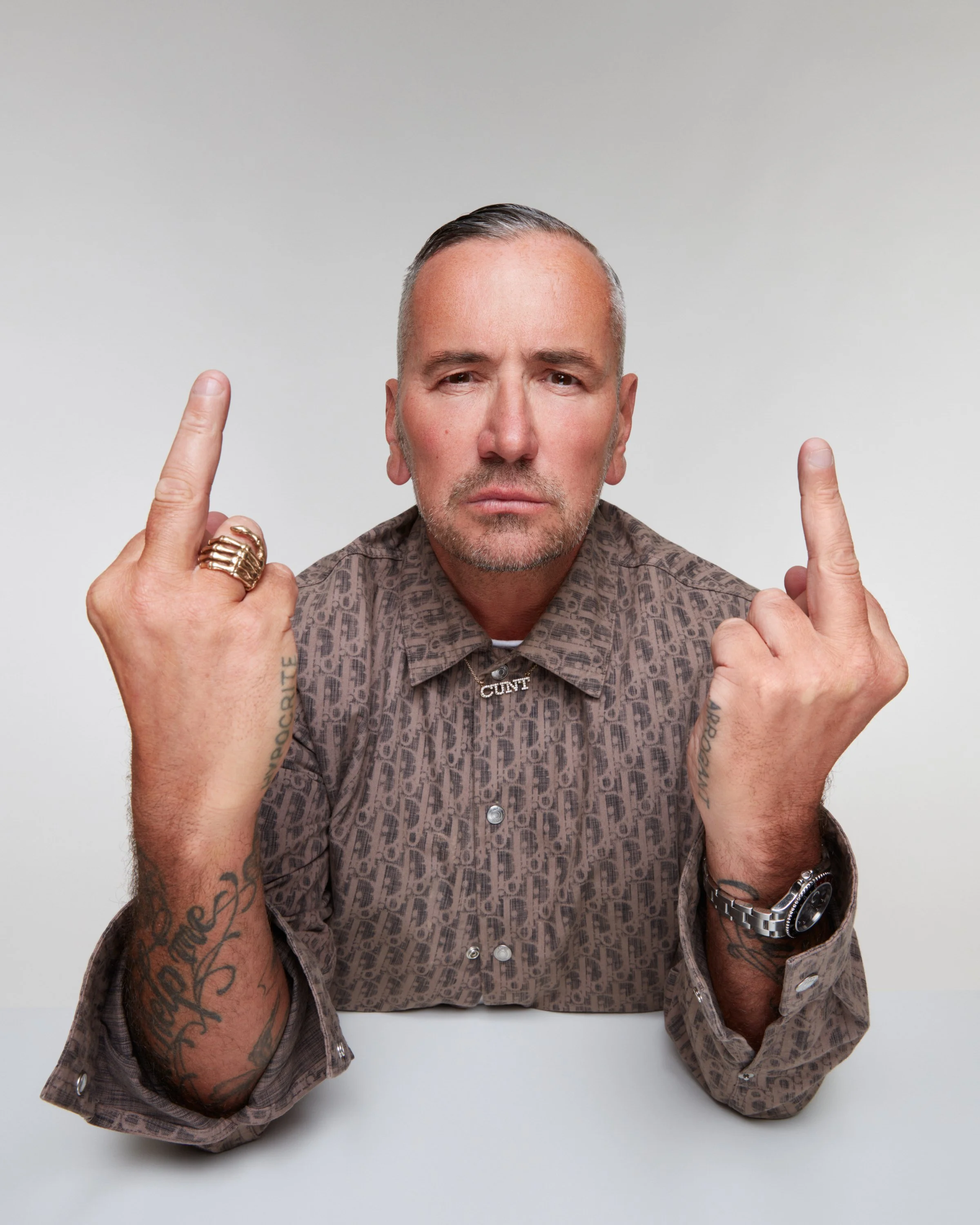 A man with tattoos on his arms and wearing a patterned shirt is making an obscene gesture with both middle fingers raised.