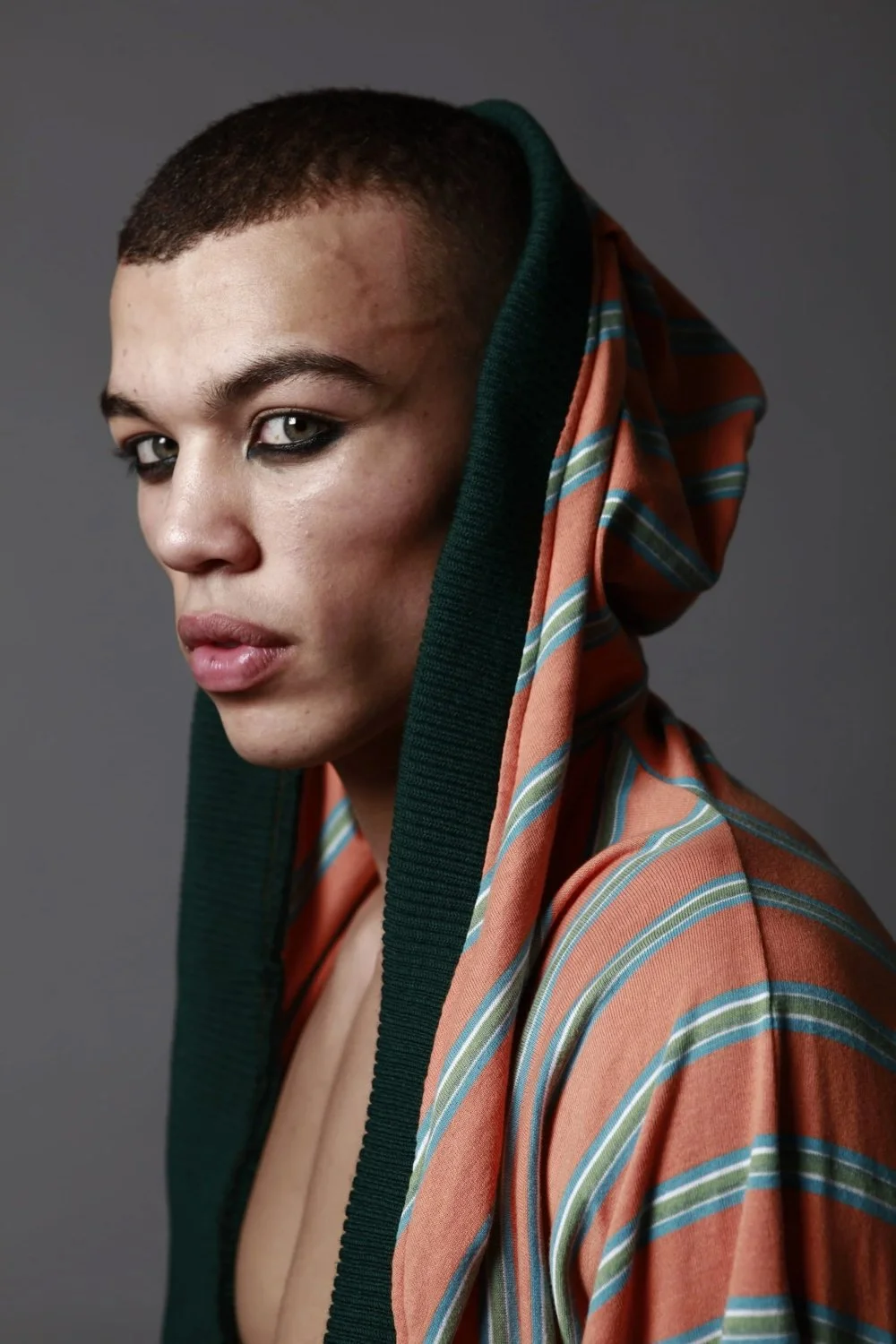 Portrait of a person with a shaved head, intense gaze, dark eye makeup, wearing a hooded, striped, orange and green jacket over a gray background.