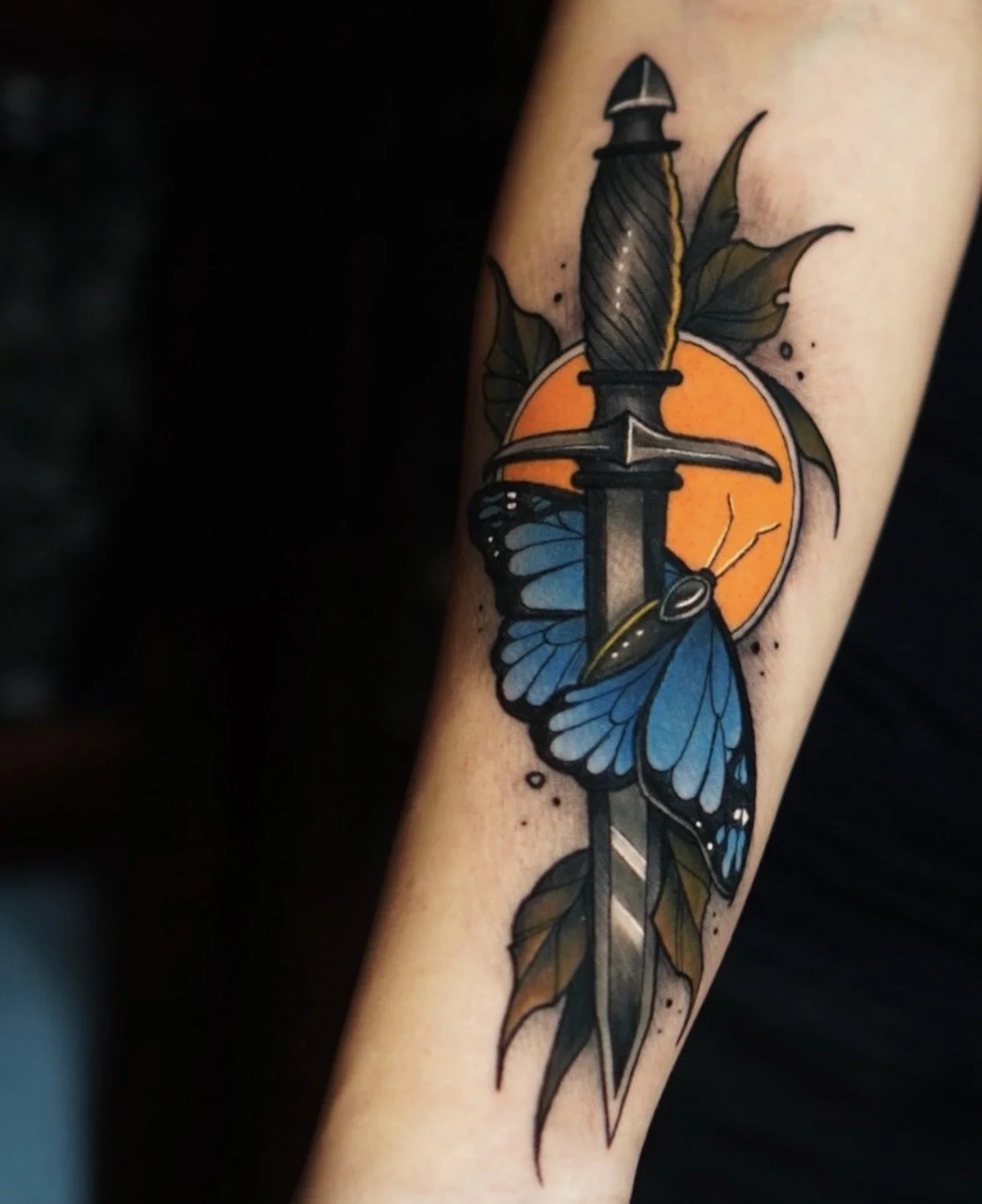 A tattoo of a dagger with a blue butterfly wrapped around it, set against an orange circle with leaves behind it.