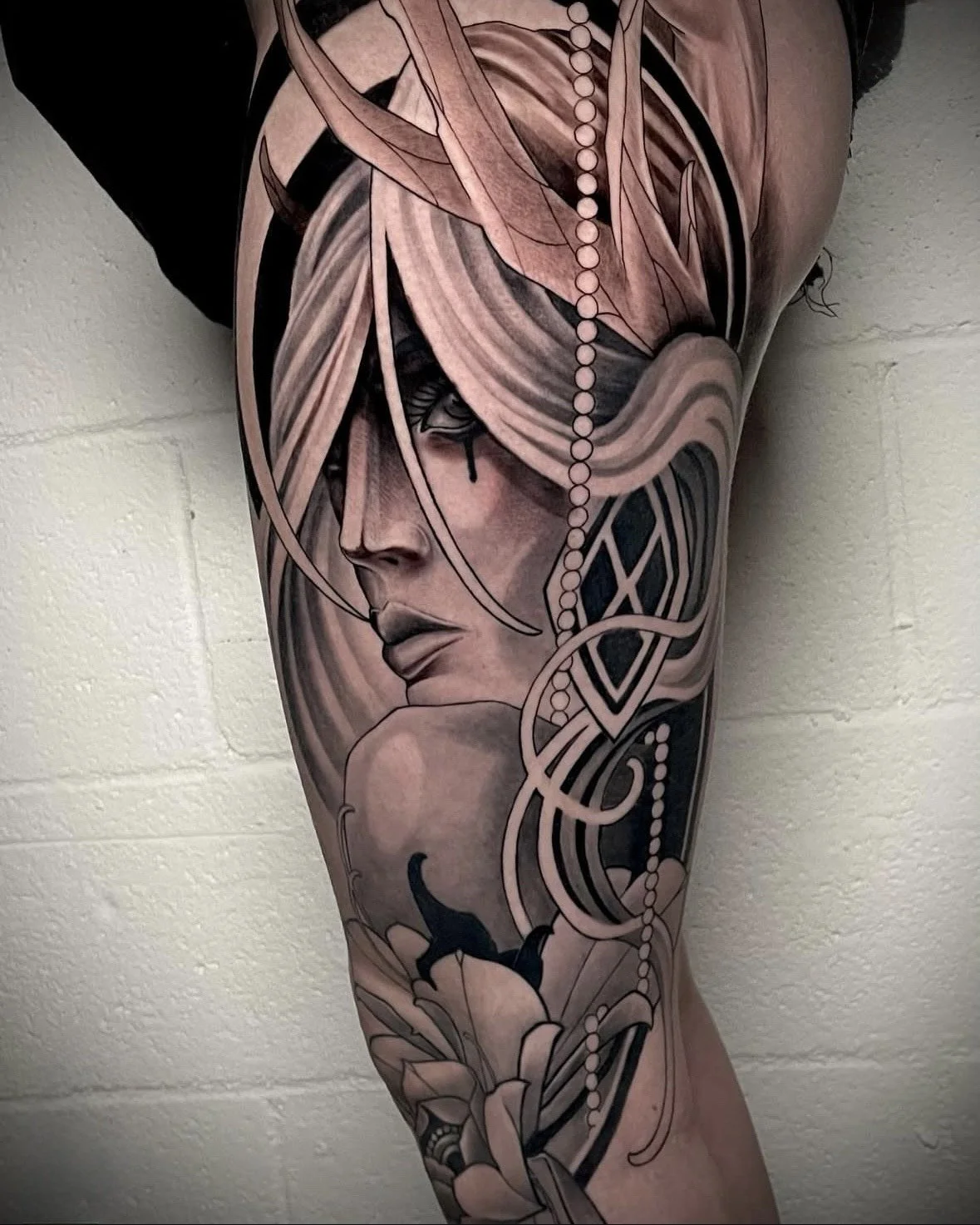 Black and grey tattoo of a woman's face with flowing hair, surrounded by decorative patterns and flowers on a person's thigh.