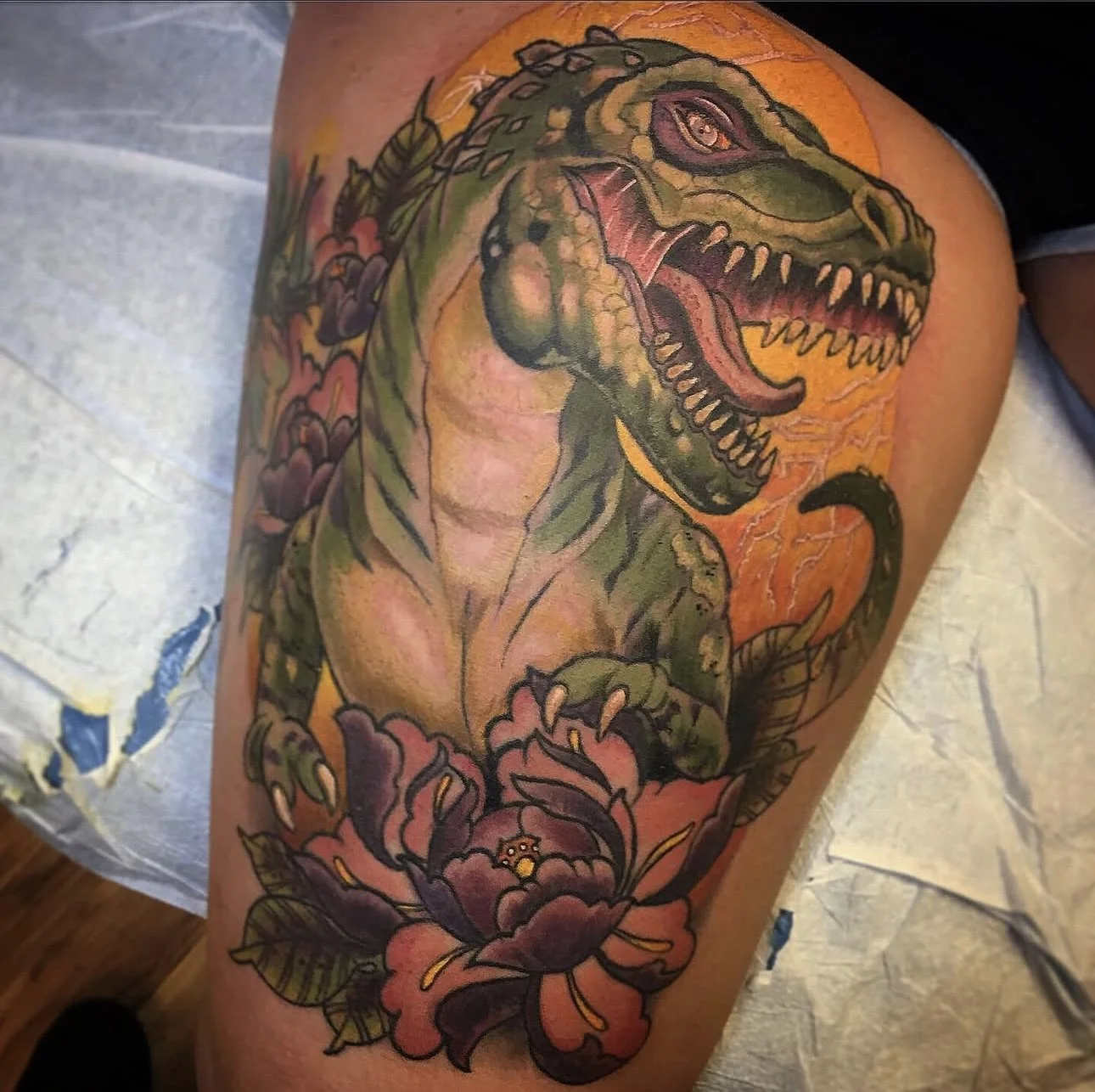 A colorful tattoo of a roaring Tyrannosaurus Rex with an orange background, green scales, and purple flowers on the limb and around the dinosaur.