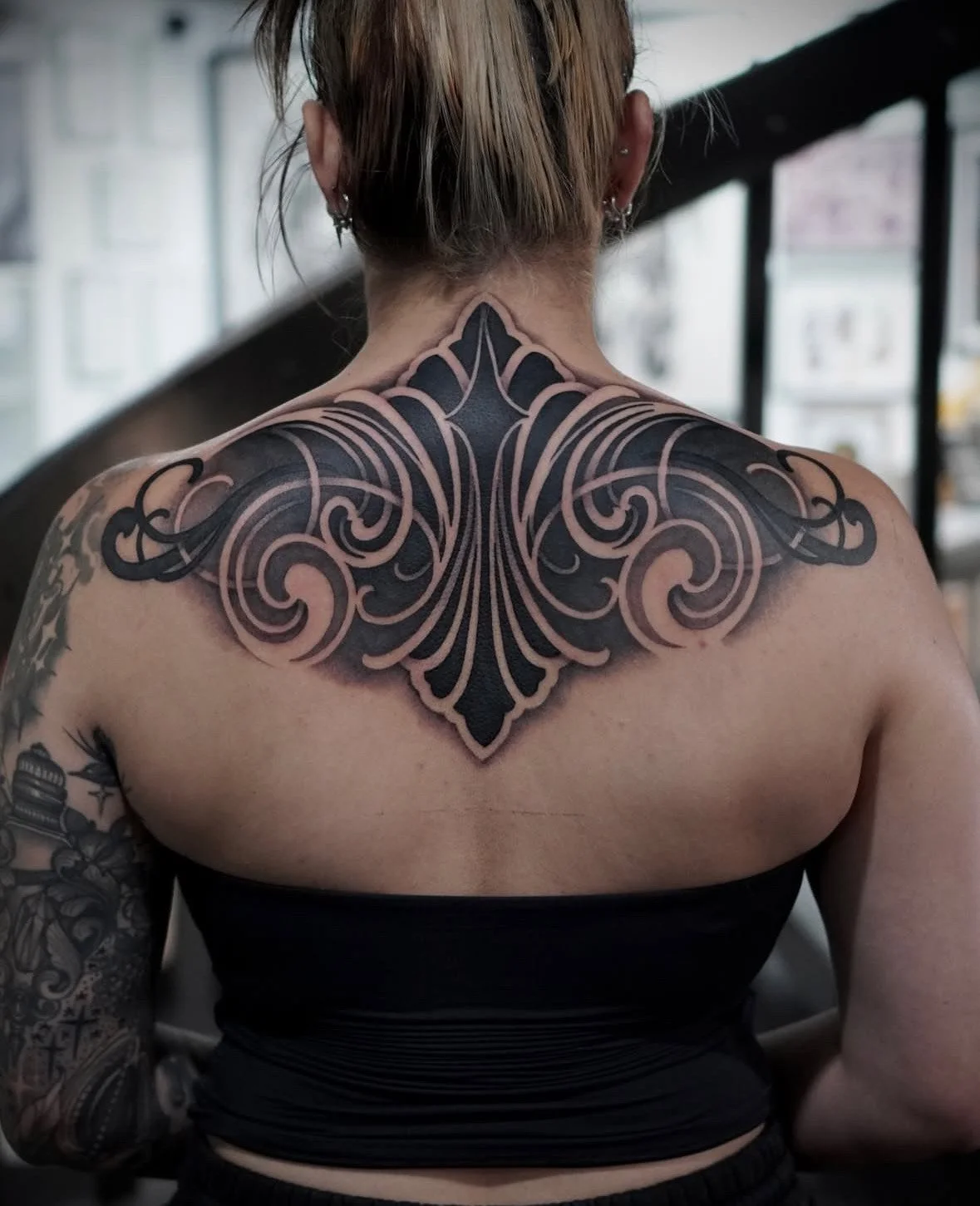 Back view of a person with a large, intricate black and gray tattoo covering the upper back and shoulders, featuring symmetrical, swirling designs and a central pointed element.