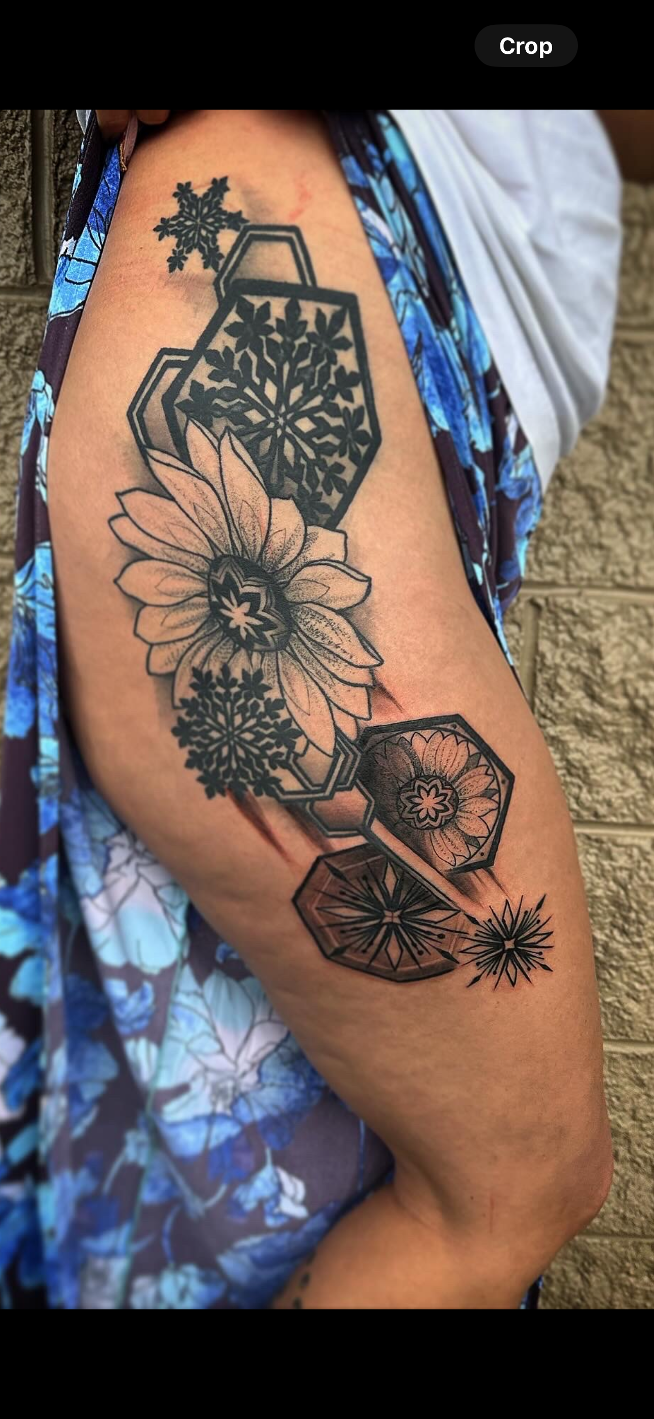 Black and white tattoo on person's arm featuring hexagonal frames with snowflakes and floral designs, including a sunflower, set against a textured beige wall background.