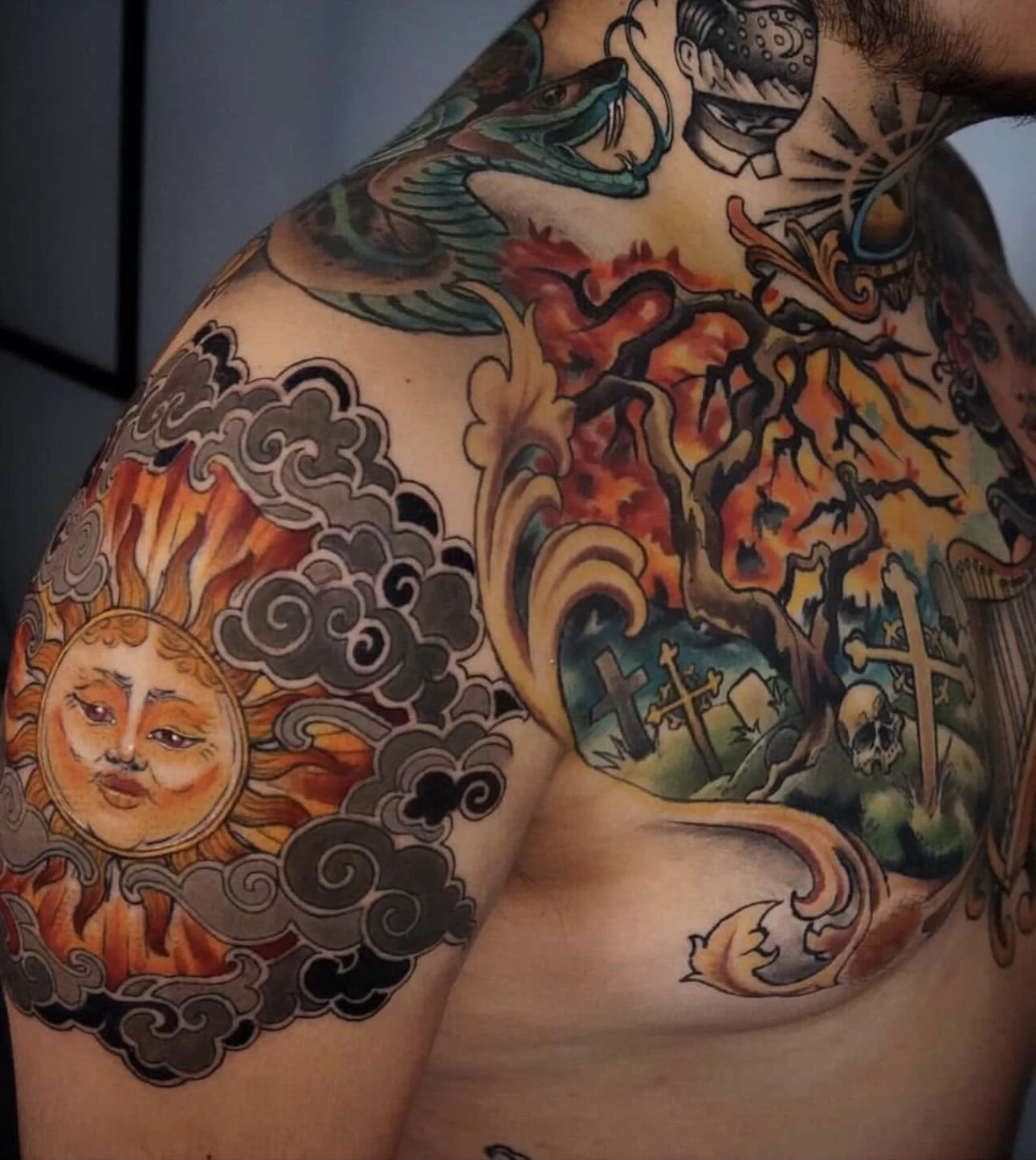 Colorful tattoo sleeve featuring a sun with a human face, swirling clouds, a dragon, a tree with candles, and crosses across the chest and shoulder.