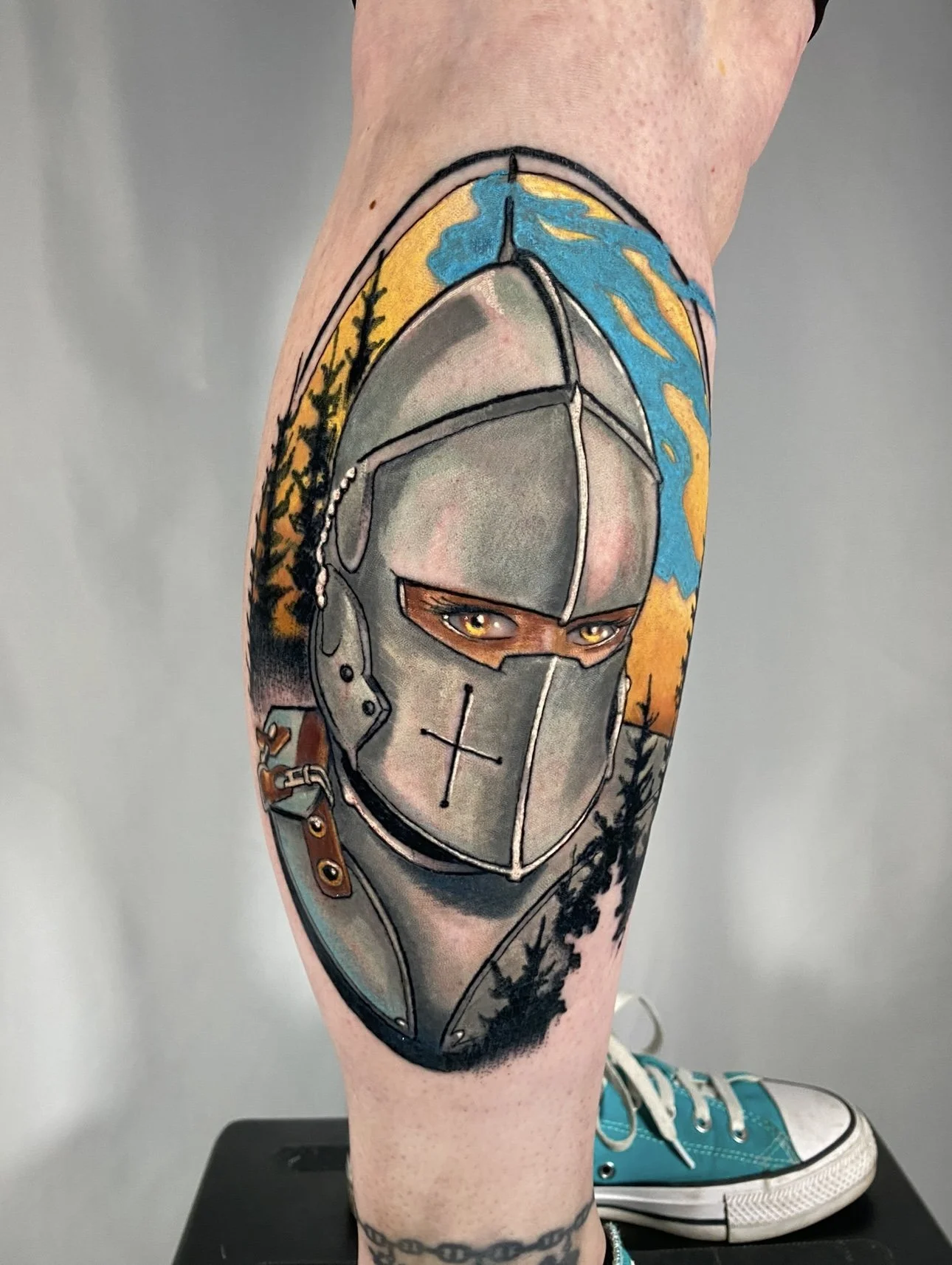 Colorful tattoo of a knight in armor with piercing eyes, set against a background of trees and a sky with blue and yellow clouds, on a person's leg.