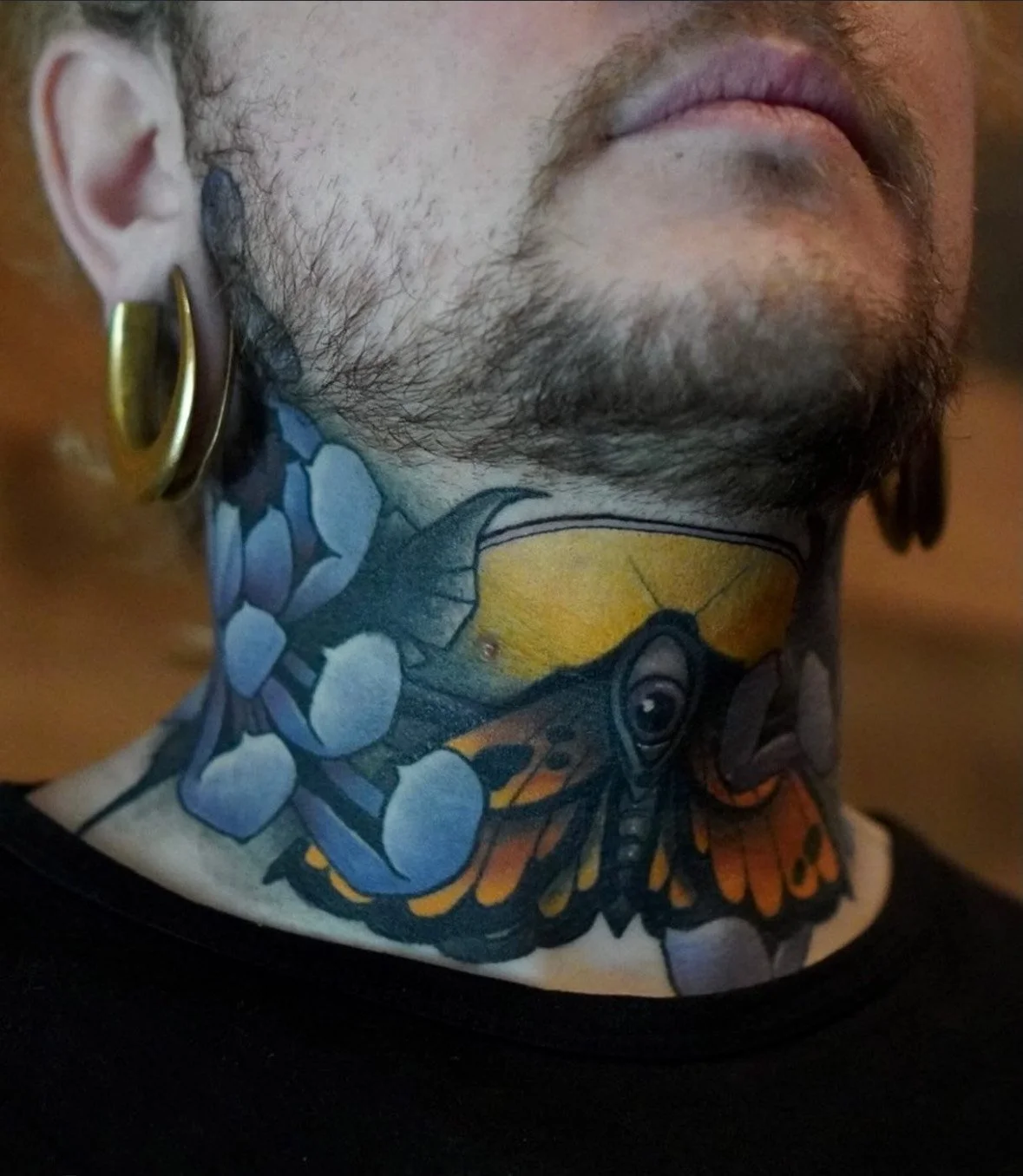 Close-up of a person's neck tattoo featuring a butterfly, blue flowers, and yellow background. They have a beard, mustache, and large gold hoop earrings.