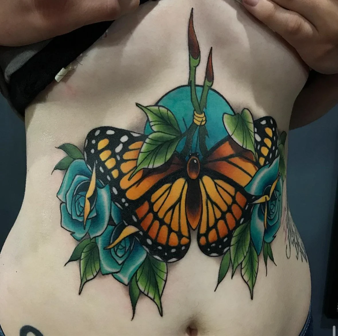 Colorful tattoo of a monarch butterfly with blue roses and green leaves on a person's chest.