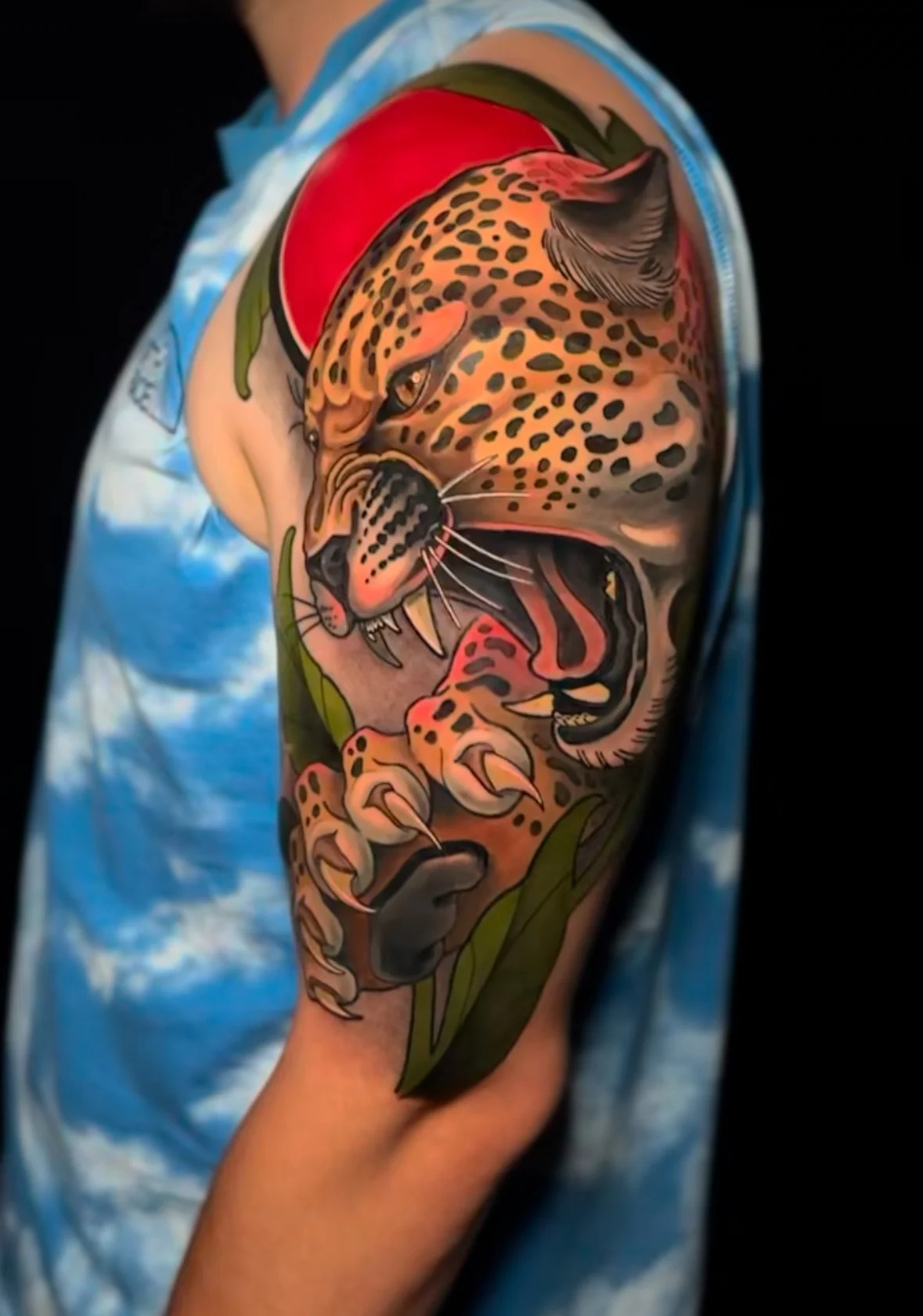 Colorful tattoo of a snarling jaguar with spots, green leaves, a red ball, and a background of blue sky with white clouds on a person's upper arm.