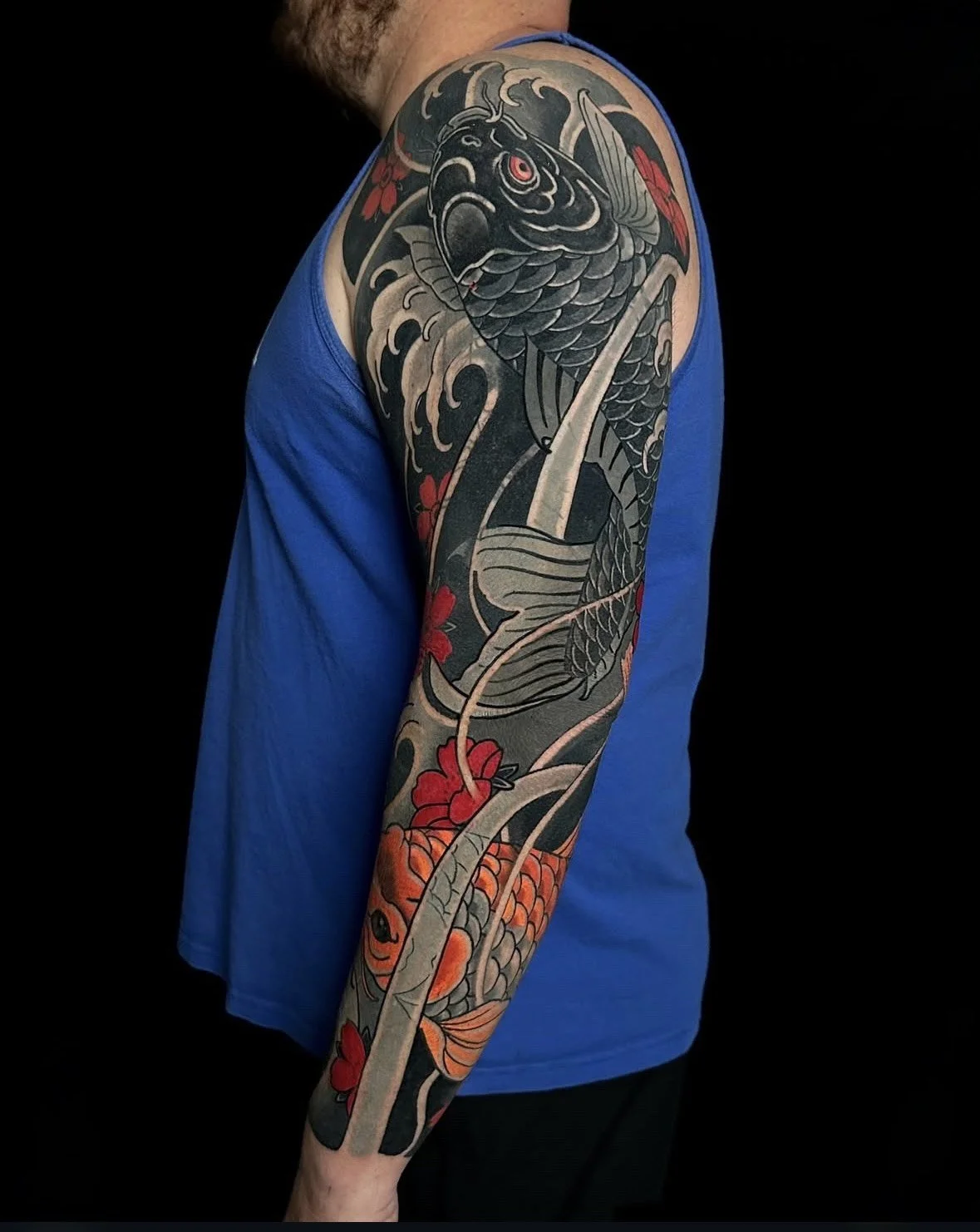 A person's arm with a detailed Japanese-style tattoo featuring a black koi fish and red flowers against a dark background.