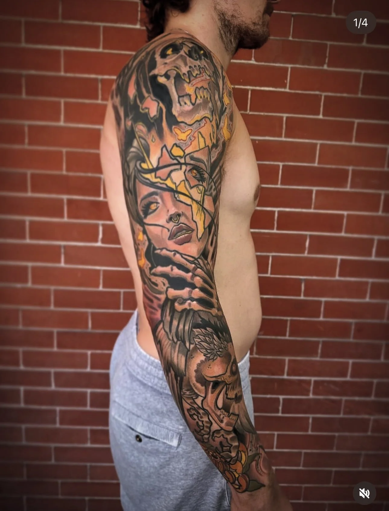 A man with a sleeve tattoo on his right arm, featuring a woman's face, a skull, a wolf, and various colorful elements, standing in front of a brick wall.