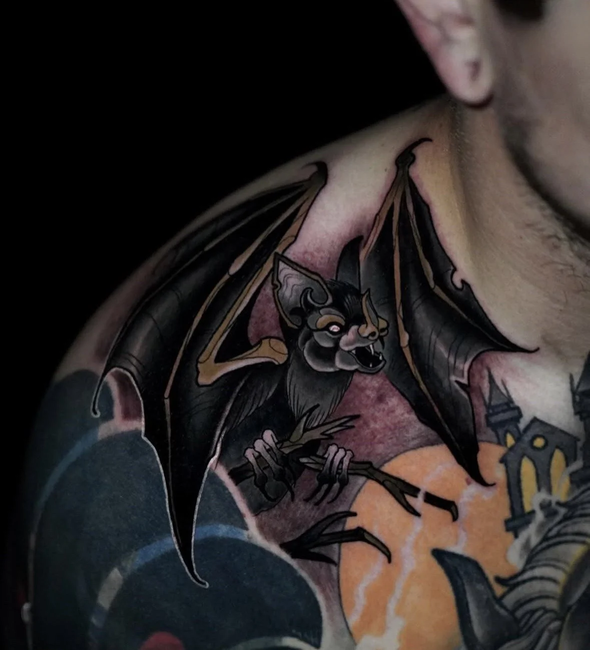 Tattoo of a black bat with a demon-like face, fangs, and wings spread, holding a small branch with leaves.