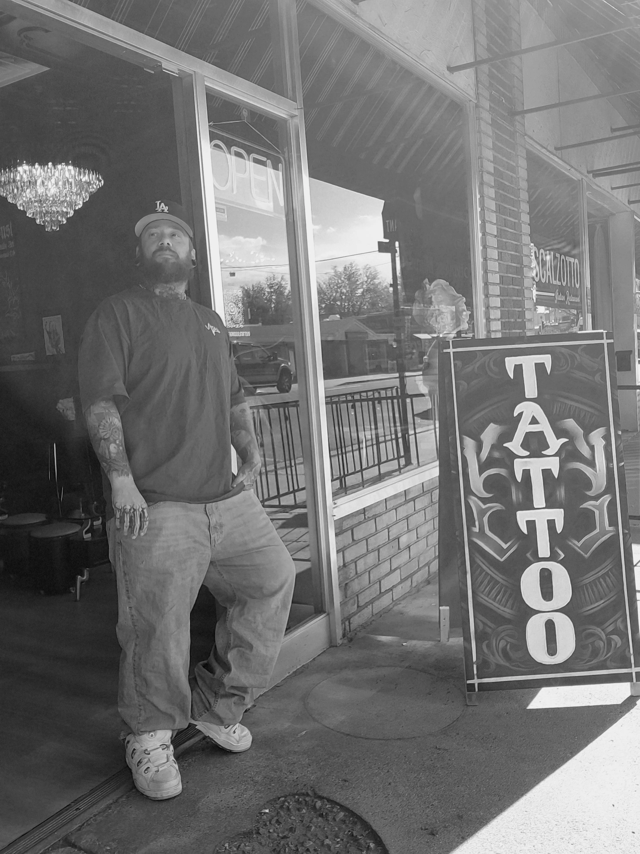 Man standing outside tattoo shop with tattoos on arms, wearing a cap, t-shirt, jeans, and sneakers, next to a sign that reads 'TATTOO' outside the shop.