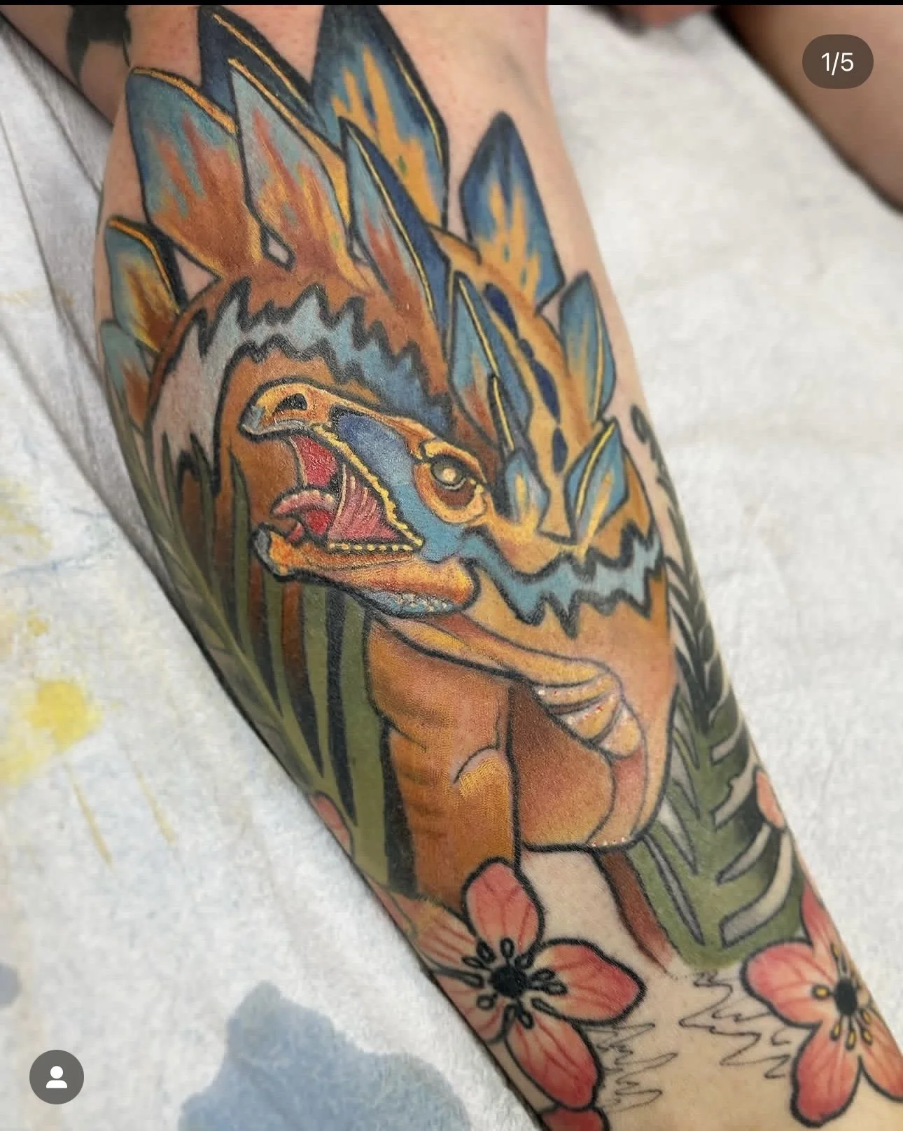 Colorful tattoo of a fierce dinosaur, possibly a T-Rex or similar theropod, with open jaws, sharp teeth, and detailed features, surrounded by floral and leafy designs.