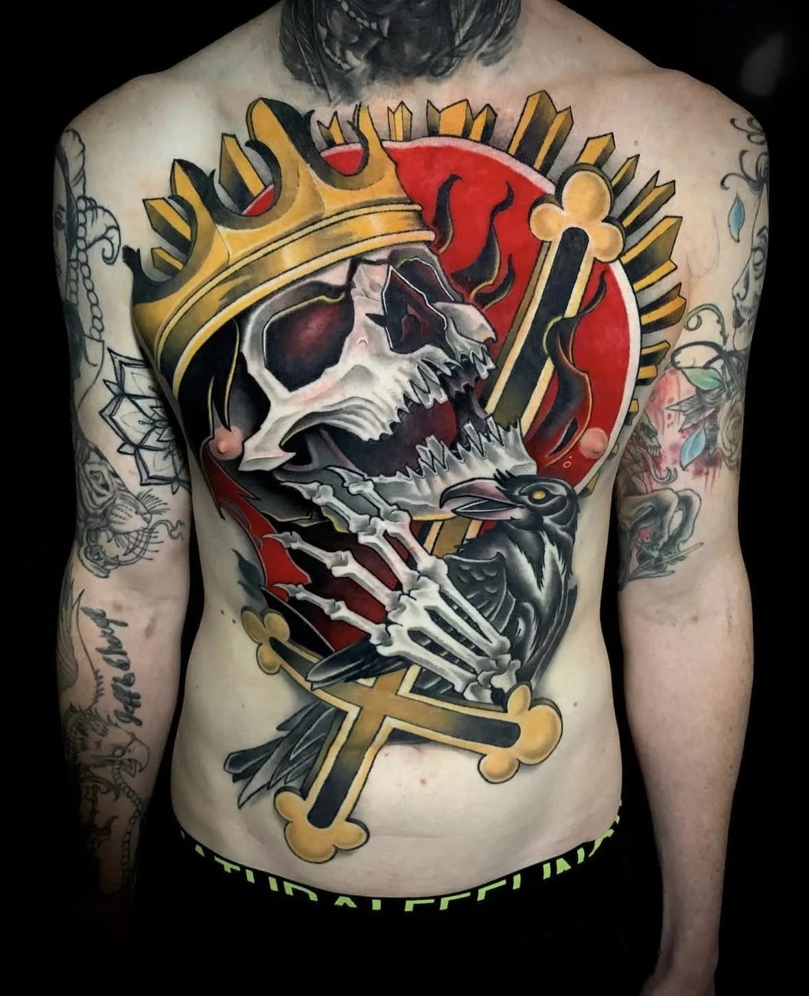 Chest tattoo of a skull wearing a crown, with a red circle background, a black rabbit holding the skull's hand, and cross-like design elements.