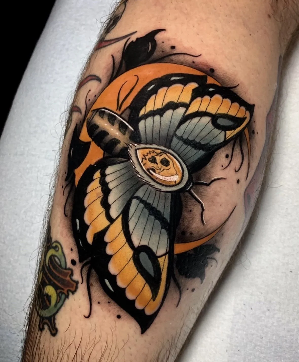 Tattoo of a butterfly with black, orange, and gray wings, featuring a skeleton face on its body and a small snake below.