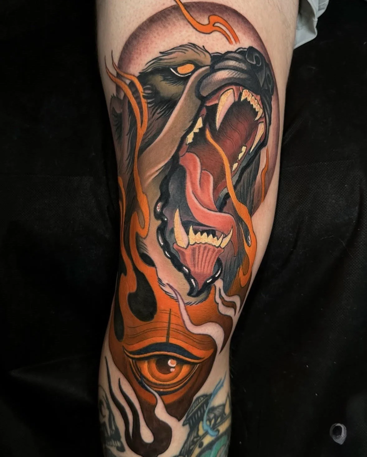 Tattoo of a roaring bear with orange flames and an eye in the lower part on a person's arm.