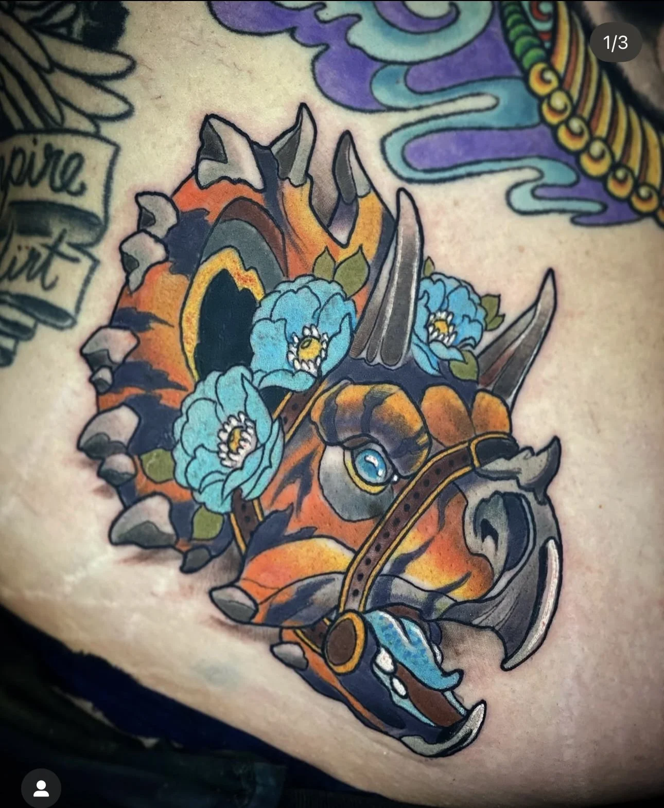 Colorful tattoo of a dinosaur skull with blue flowers and orange, black, and gray details.