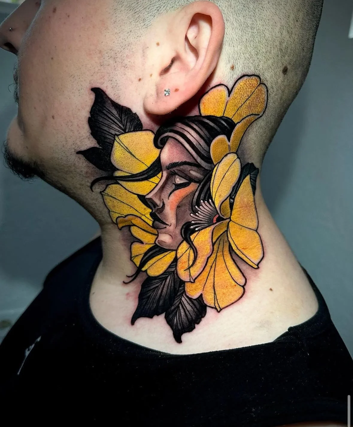Colorful tattoo of a woman's face, surrounded by yellow butterfly wings and black leaves, on the side of the neck.
