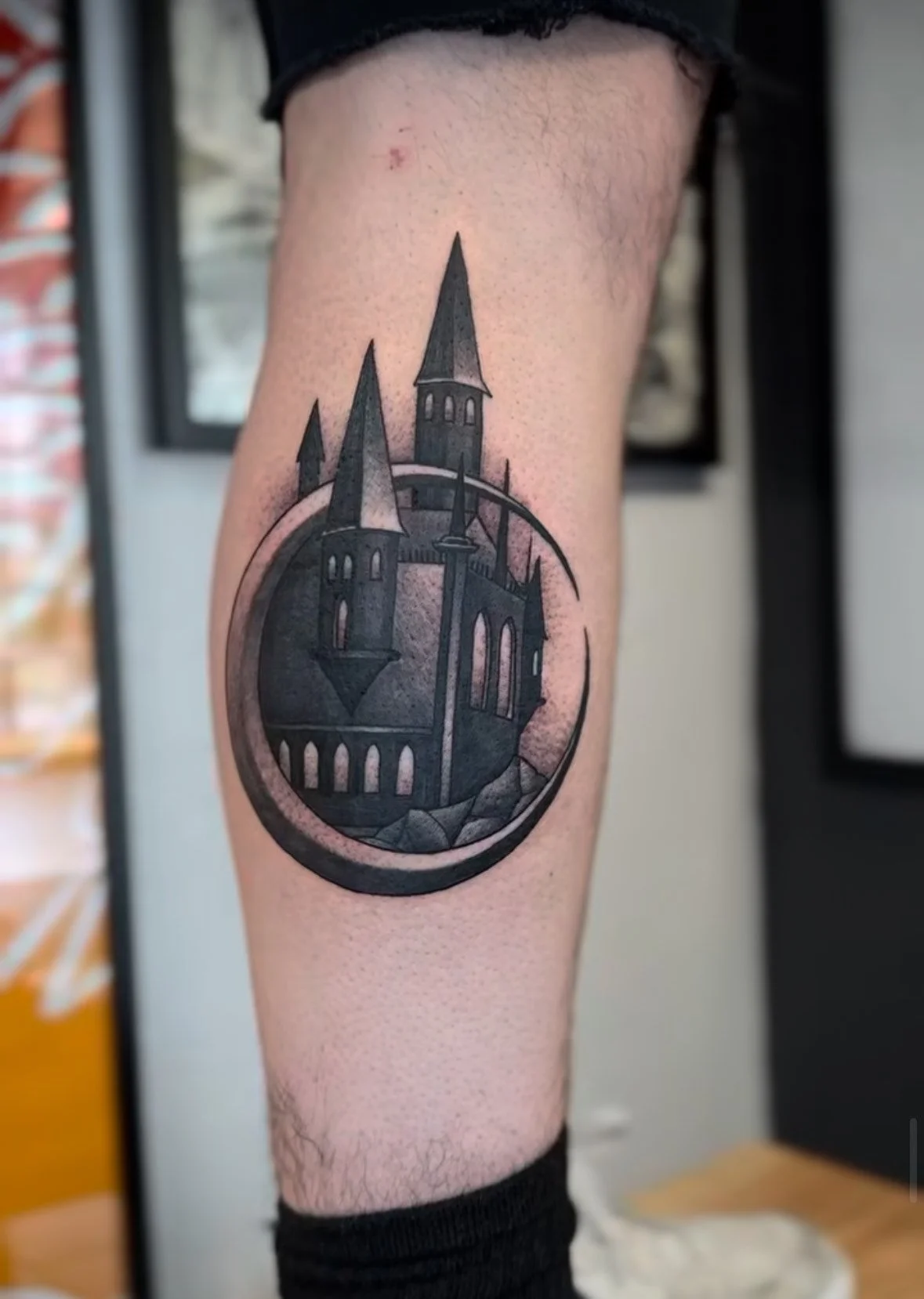 Tattoo of a dark, gothic castle with tall spires, enclosed within a circle, on a person's thigh.