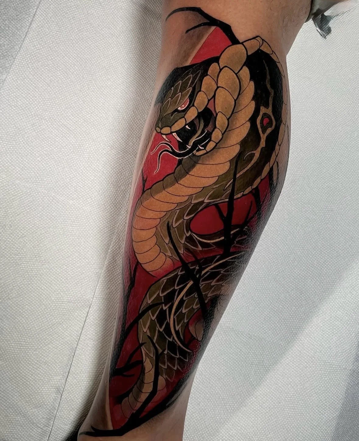 Tattoo of a snake with its mouth open, detailed scales, and red eyes on a person's leg, with a red background.