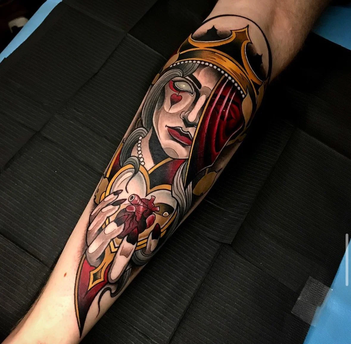 Tattoo of a woman with long hair, pearls, and red lips holding a human heart, with a crown and red drapery in the background.
