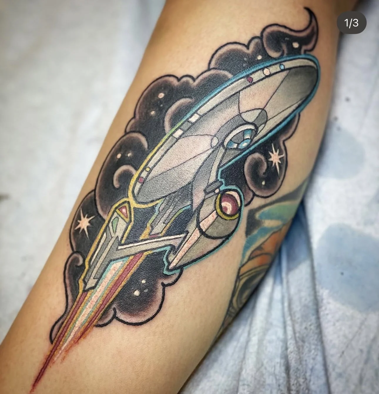 Tattoo of a spaceship with vibrant colors, surrounded by black clouds and stars.