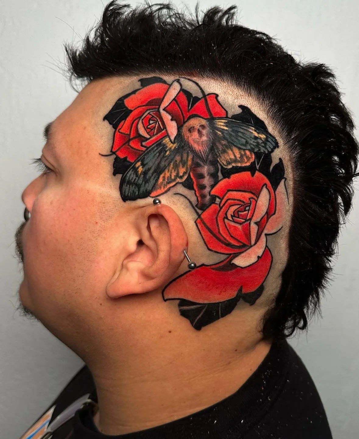 A person with a colorful tattoo of a moth and roses on the side of their head, and with ear and nose piercings.