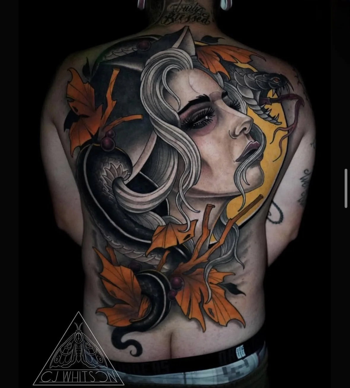 A detailed tattoo covering the person's back, featuring a woman's face with flowing hair, a snake, autumn leaves, and a crescent moon in a dark background.