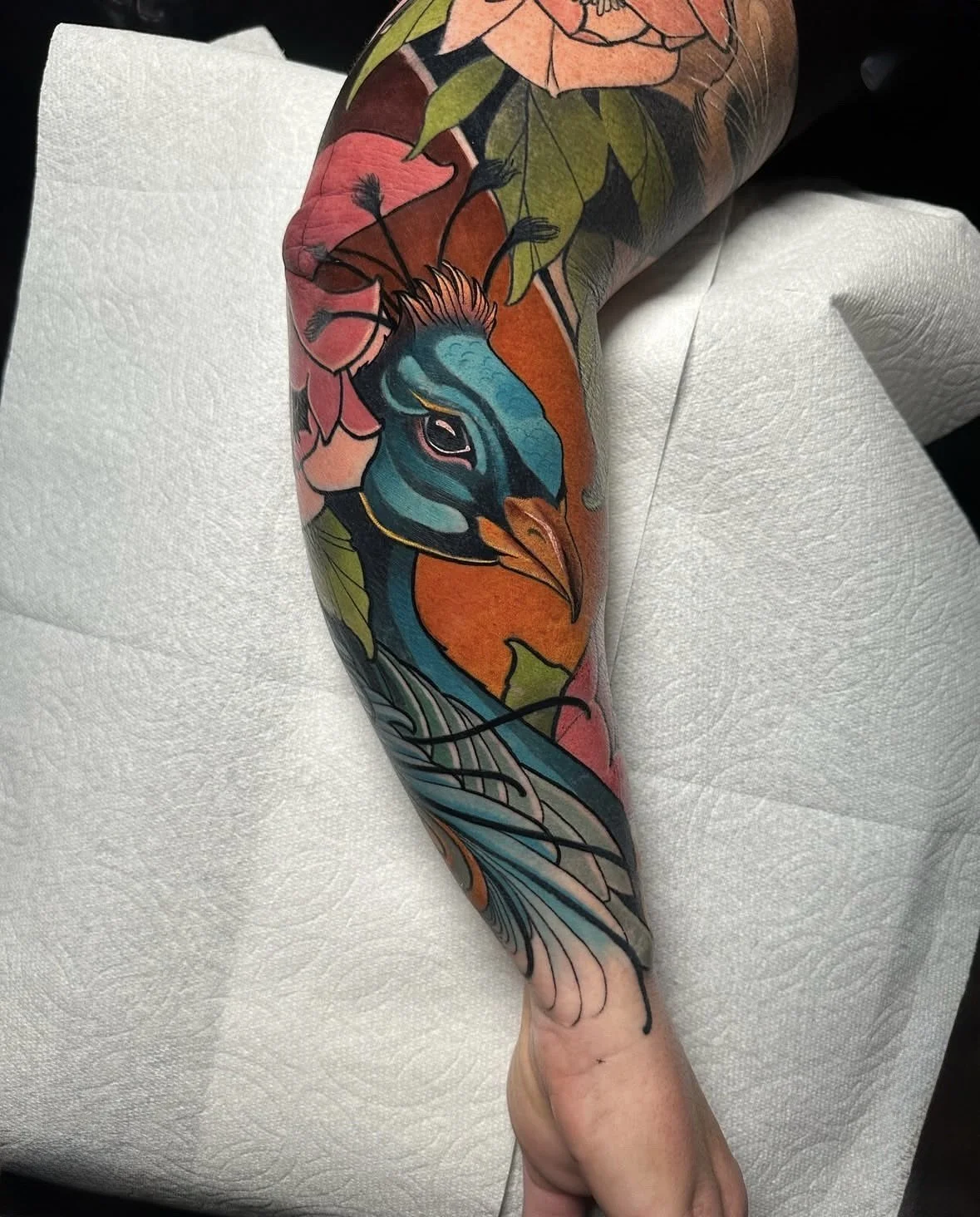Colorful tattoo of a peacock with pink flowers and green leaves on person's arm.