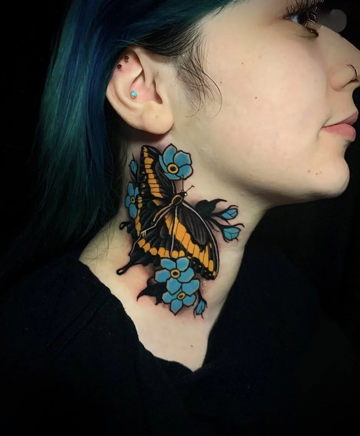A woman with blue-green hair, multiple earrings, and a septum piercing shows a colorful tattoo of a butterfly and blue flowers on her neck.