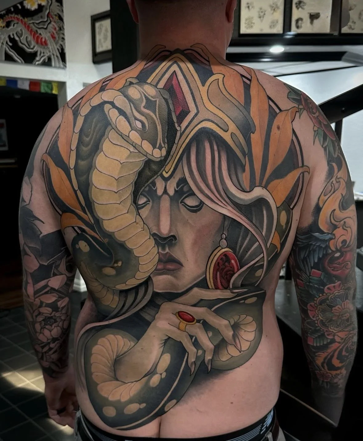 Tattoo of a woman's face with closed eyes, adorned with earrings, with a snake wrapped around her head and arm, featuring floral designs on the arms.