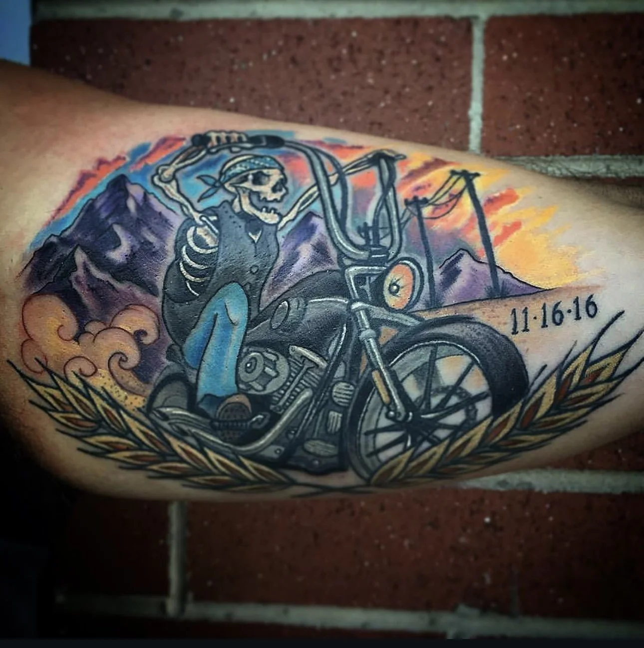 Tattoo of a skeleton riding a motorcycle with mountains and sunset background, date '11-16-16', and decorative laurel leaves.