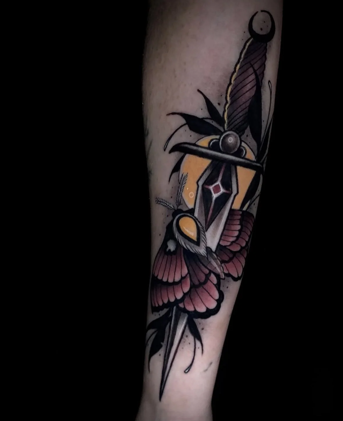 Tattoo of a dagger piercing a butterfly with a moth, surrounded by black leaves and yellow accent shapes, on the person's forearm.
