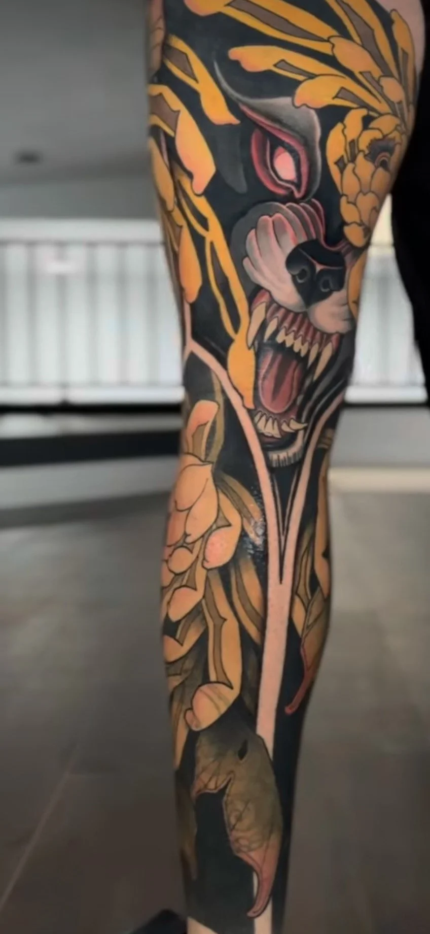 Tattoo of a fierce lion's face with sharp teeth, surrounded by orange and yellow floral patterns on a person's leg.