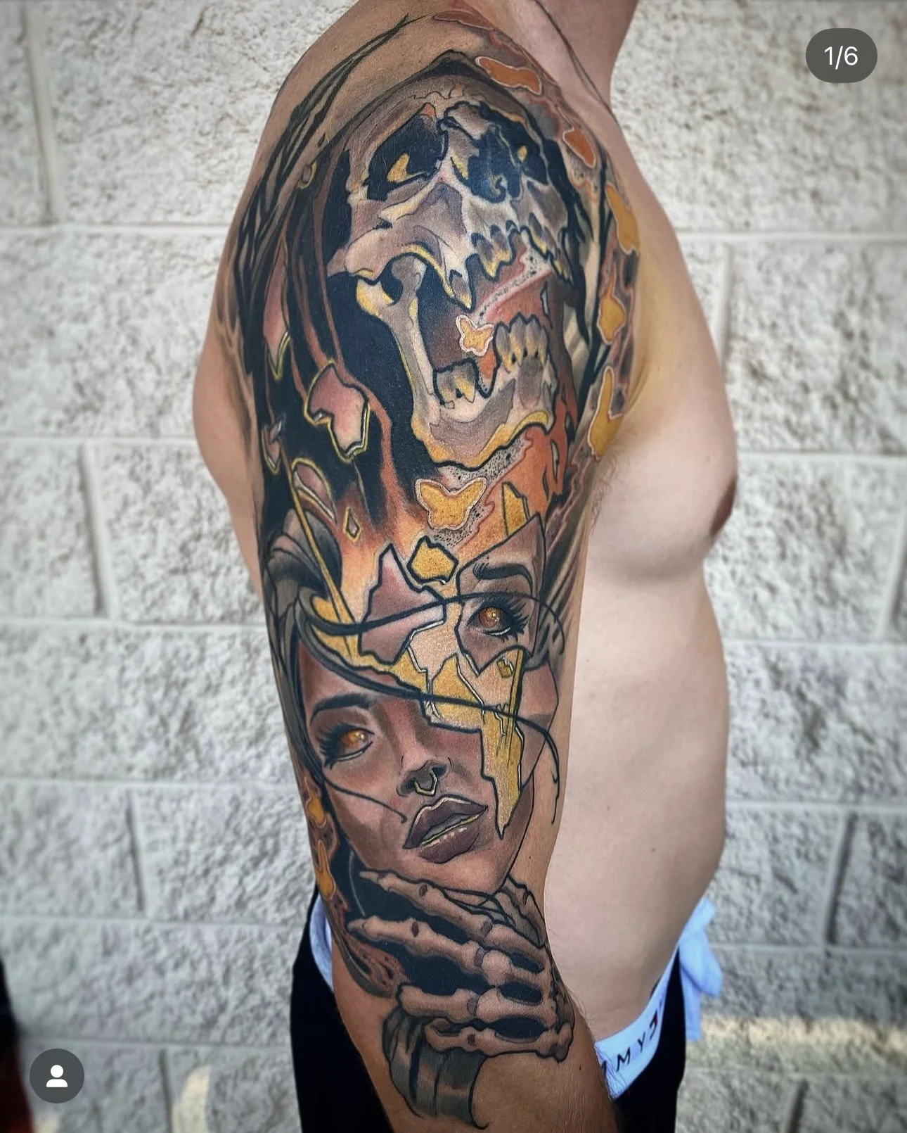 Tattoo of a woman's face with black hair and golden eyes on a person's upper arm. Above the face, there is a skull with an open mouth, and a skeleton hand is near the face holding a painting brush, all in a colorful, abstract style.