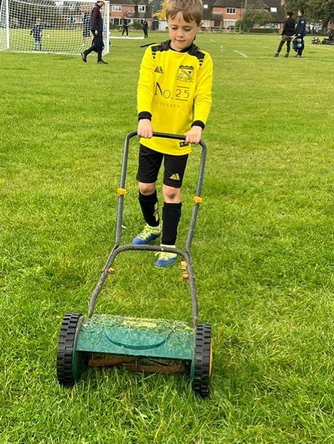 A young boy in a yellow sports jersey and black shorts is pushing a grass roller on a soccer field. There are people in the background near a goalpost and some houses.