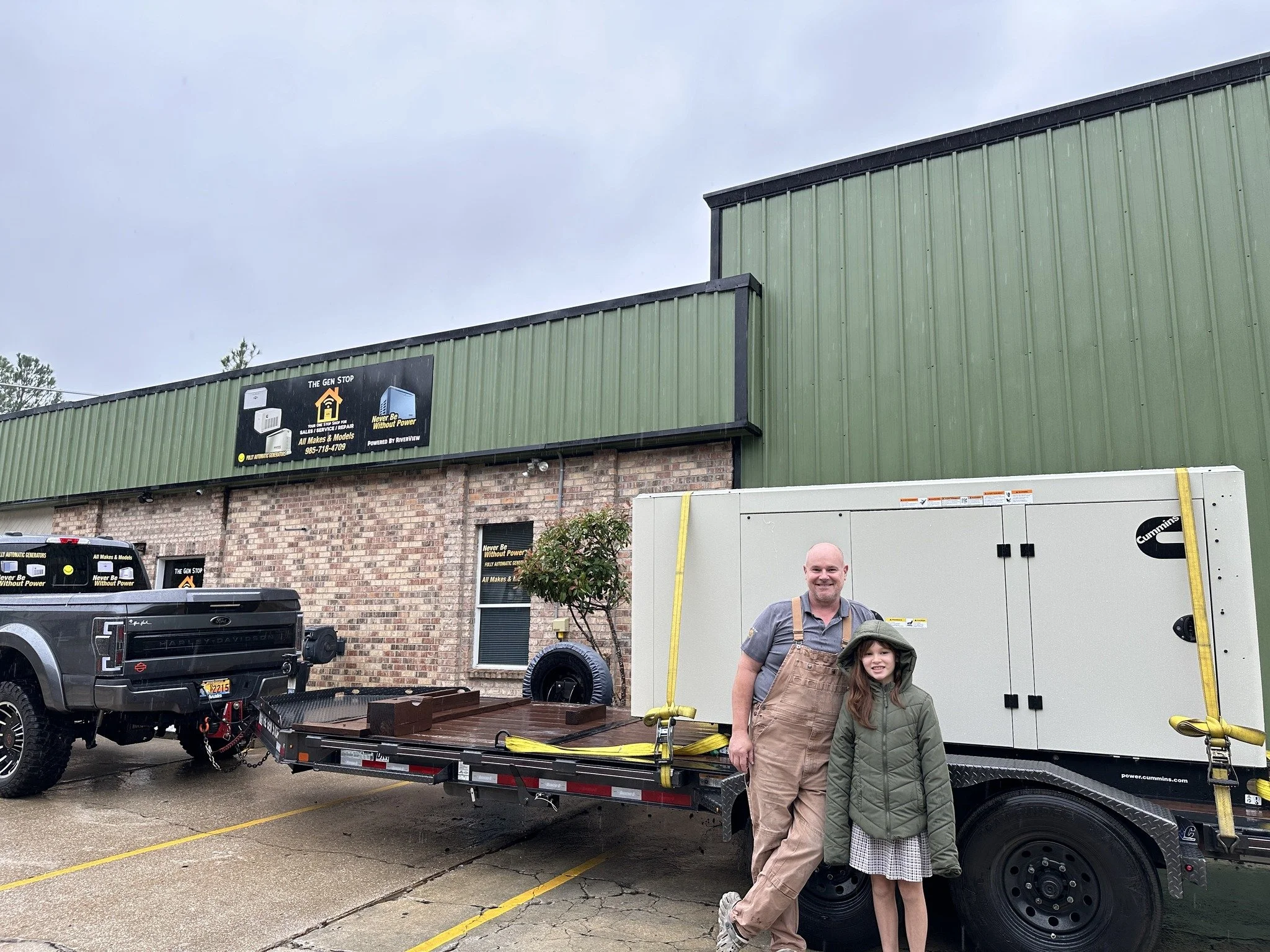 A man and a young girl standing in front of a large mobile generator on a trailer, parked outside a commercial building with a green metal facade and brick lower wall, on a rainy day.