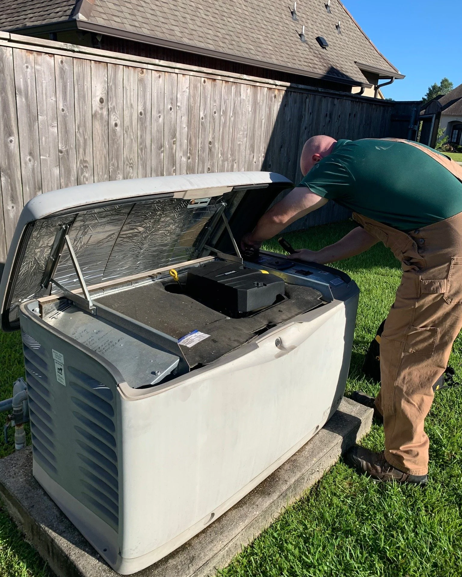 A man repairing or servicing the inside of a white residential air conditioning unit on a concrete slab outside a house, with a wooden fence and house roof in the background.
