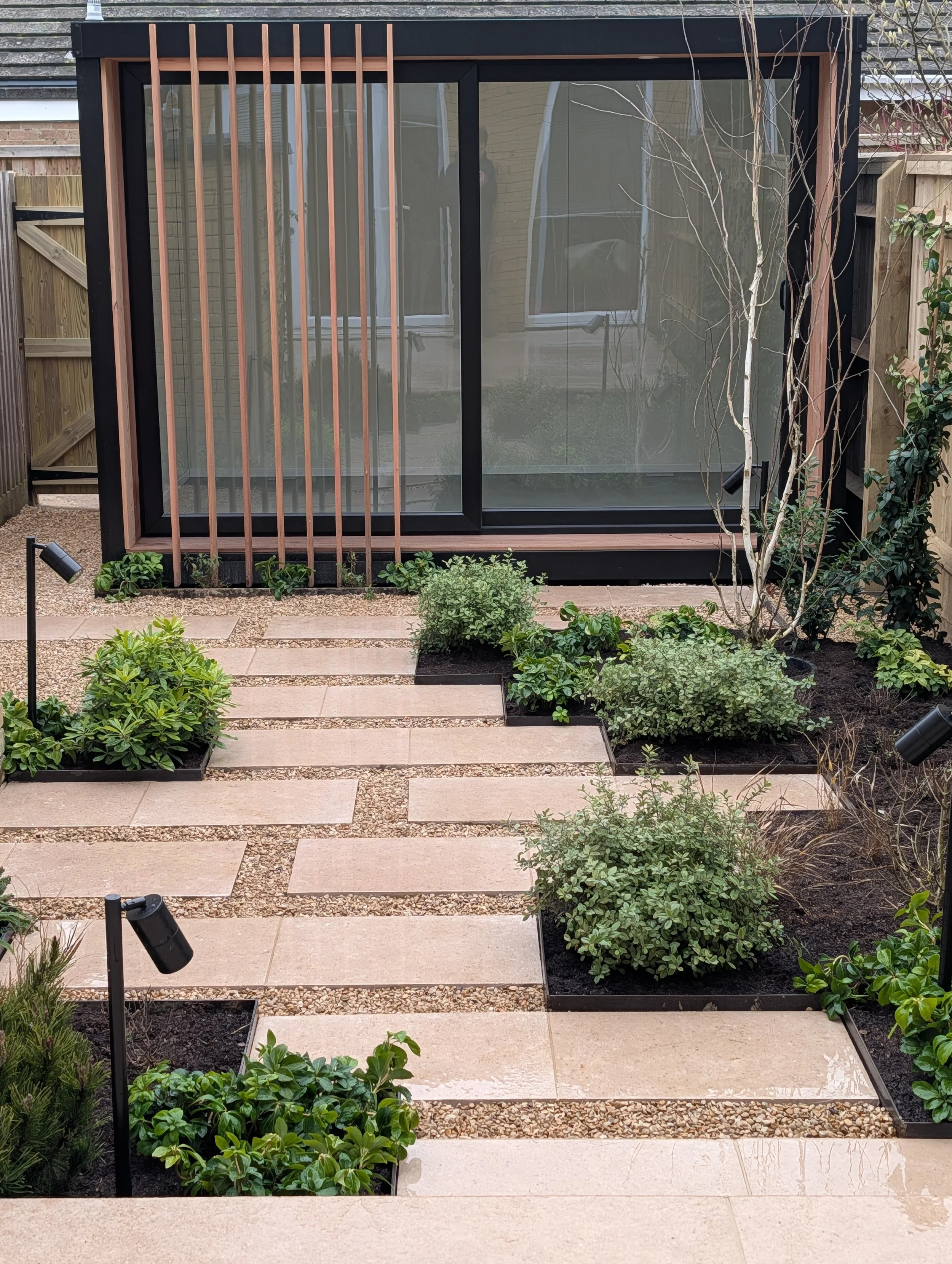 A modern backyard garden with a black-framed glass structure, potted plants, shrubs, pathway stones, and small outdoor lights.