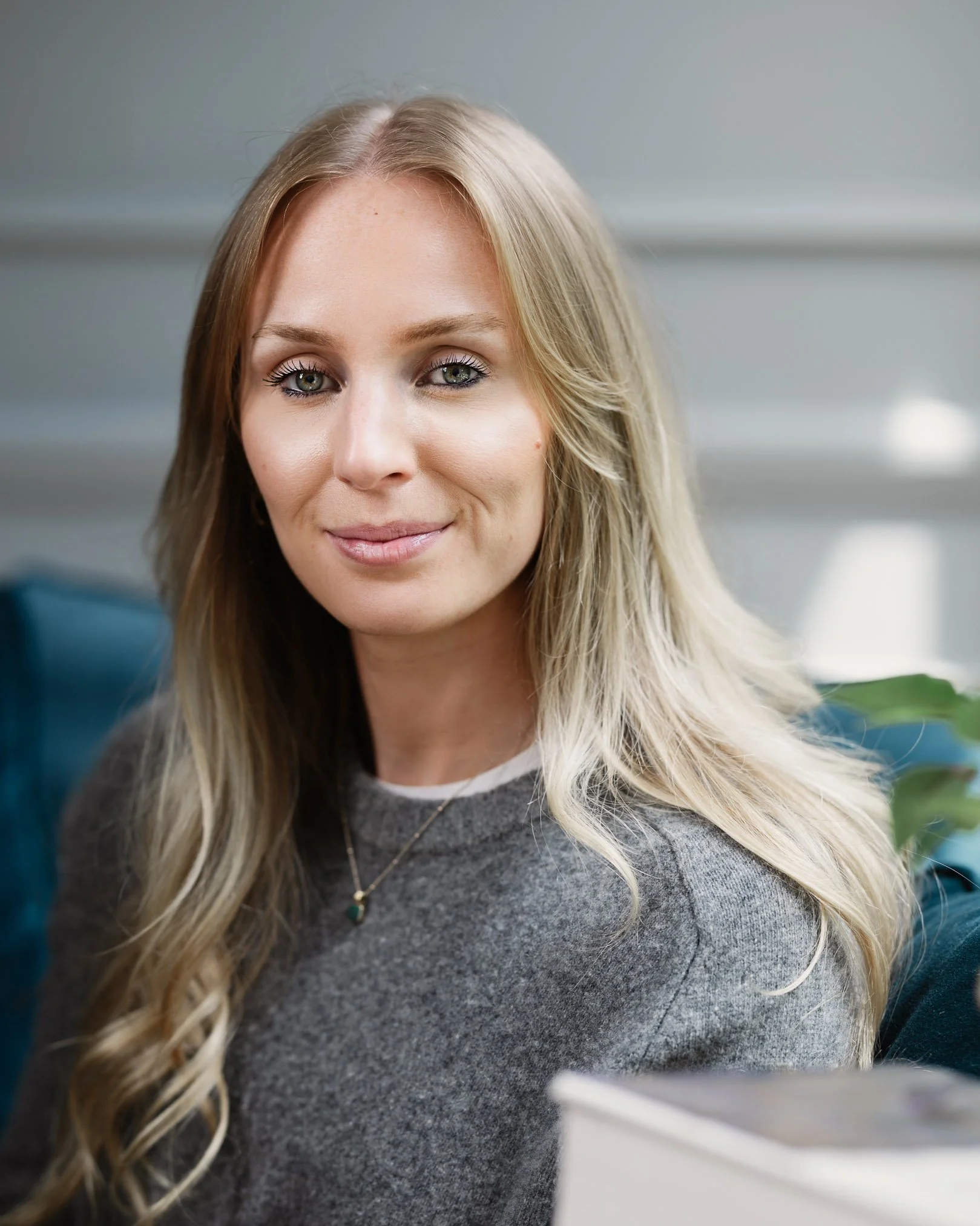 A woman with long blonde hair and blue eyes sitting on a blue couch, smiling softly at the camera in a cozy indoor setting.