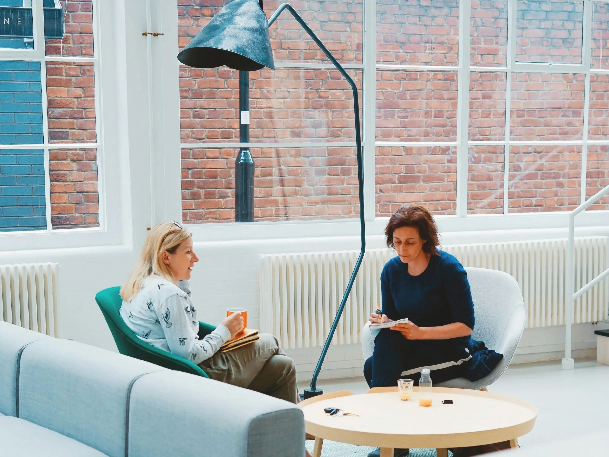 Two people having a conversation in a private space. One of the women is holding a book to refer to the conversation. The other is holding a coffee mug while listening