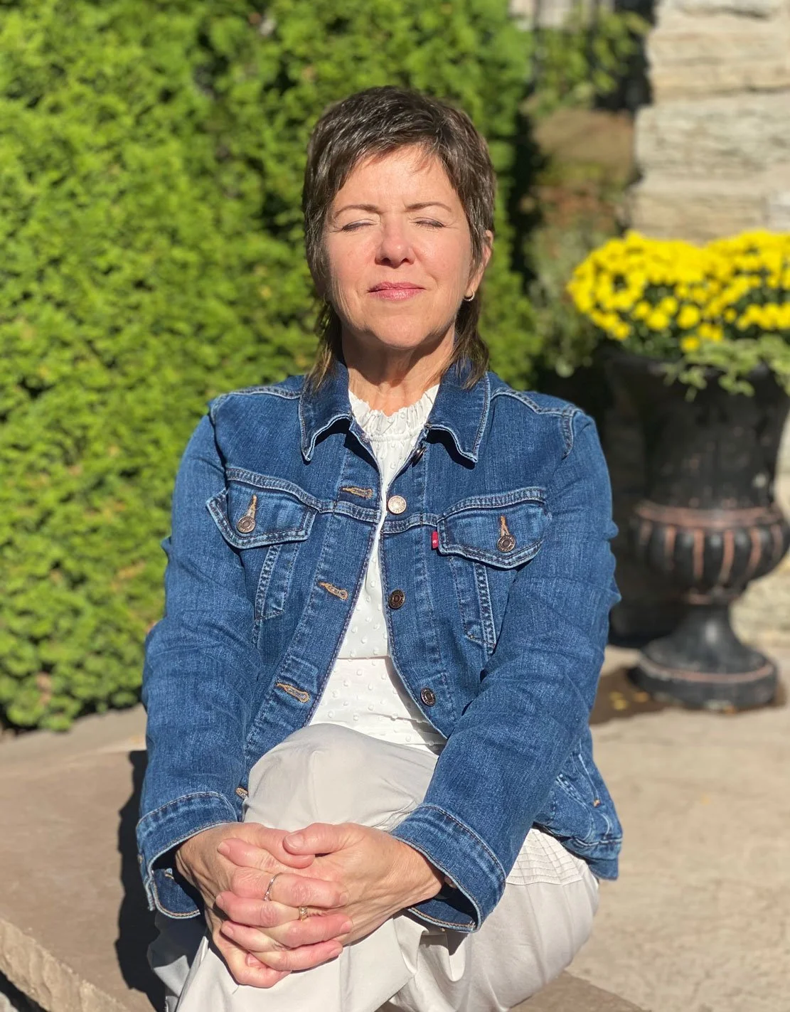 Woman sitting outdoors in sunlight with eyes closed, wearing a denim jacket and light-colored clothing, hands resting in her lap, surrounded by greenery.
