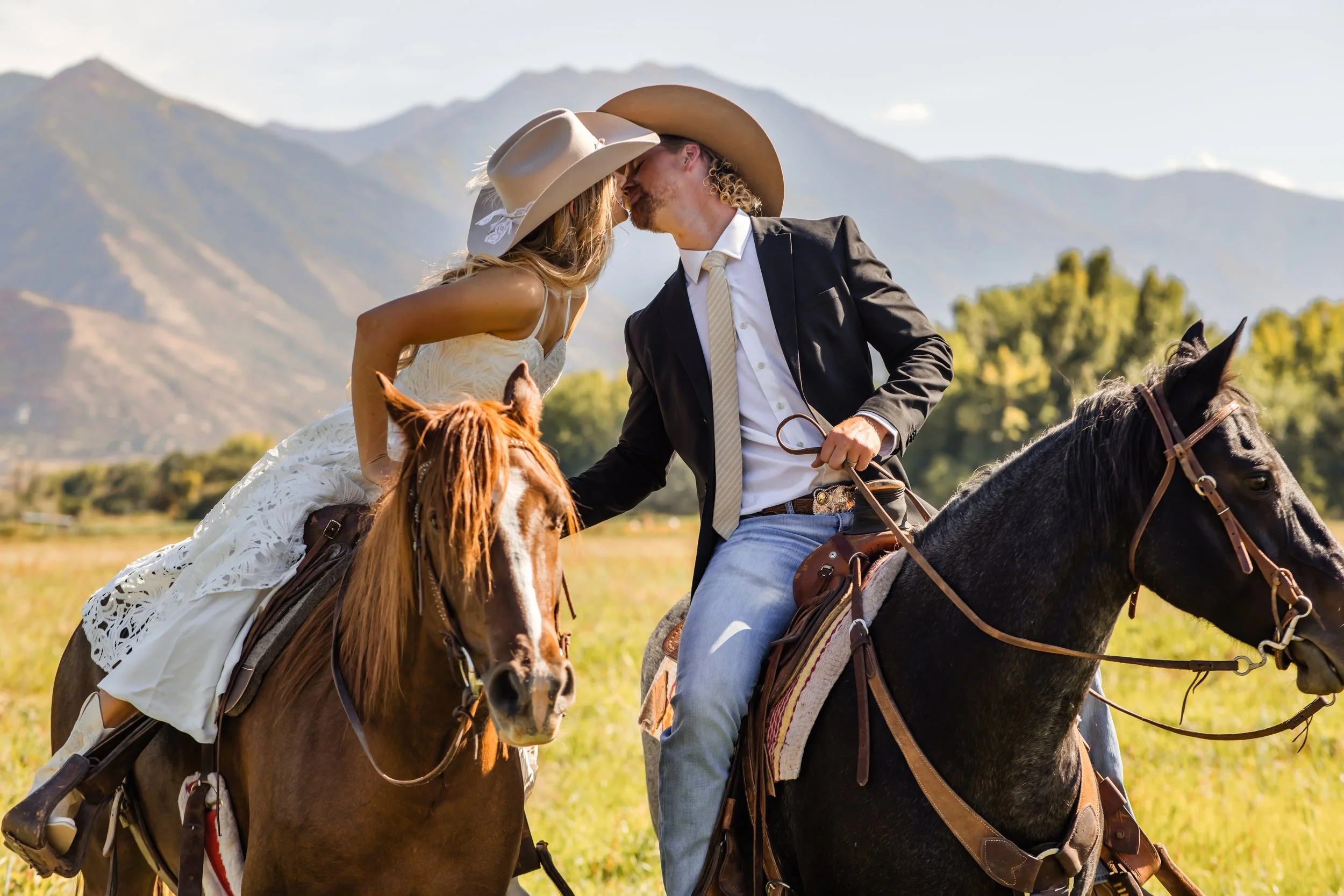 A man and woman kissing while riding horses outdoors with mountains in the background, both wearing cowboy hats.