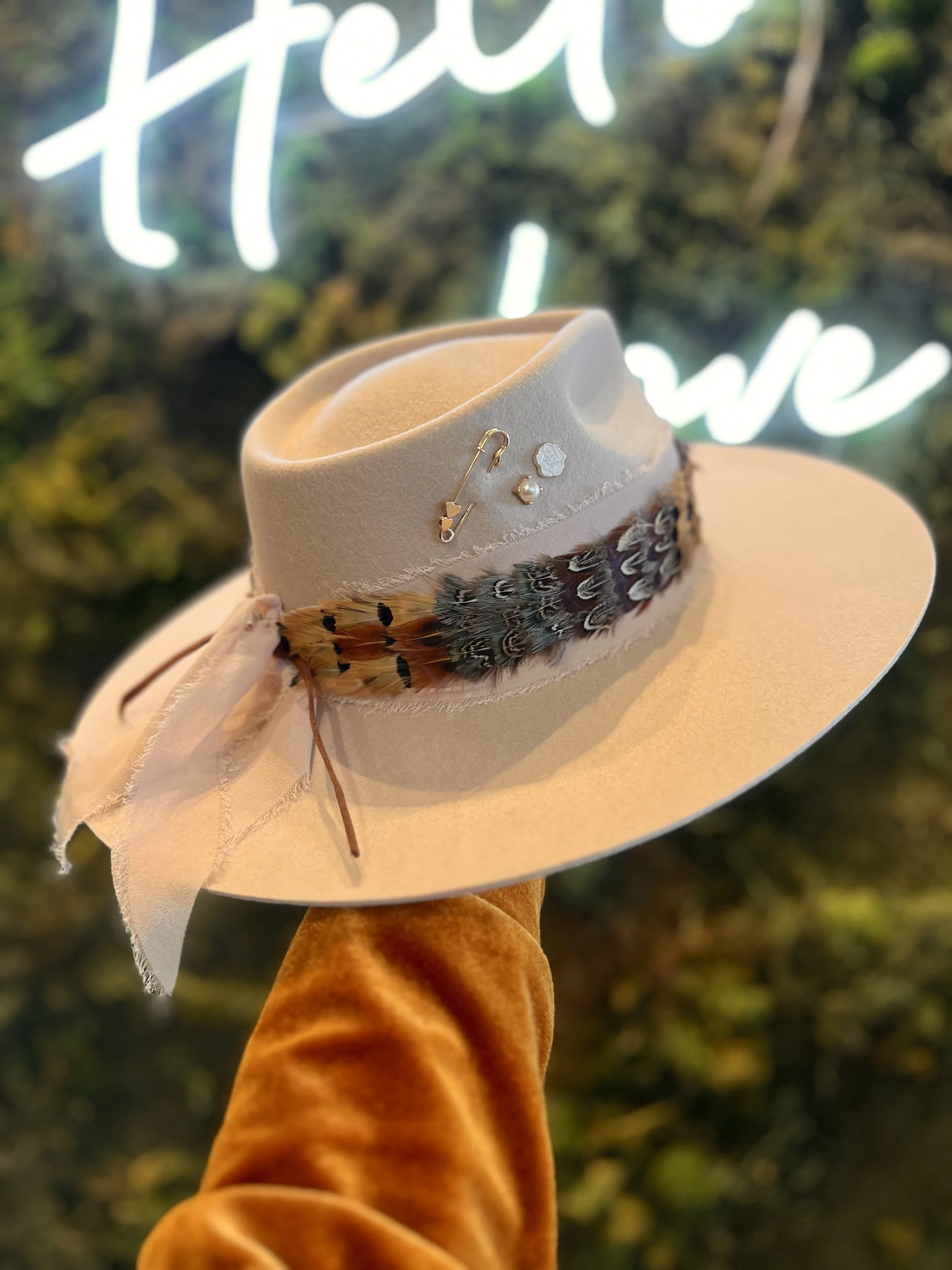 A beige wide-brimmed hat adorned with feathers and jewelry, held up by a person wearing an orange velvet sleeve, with a blurry green and white neon sign reading 'free' in the background.
